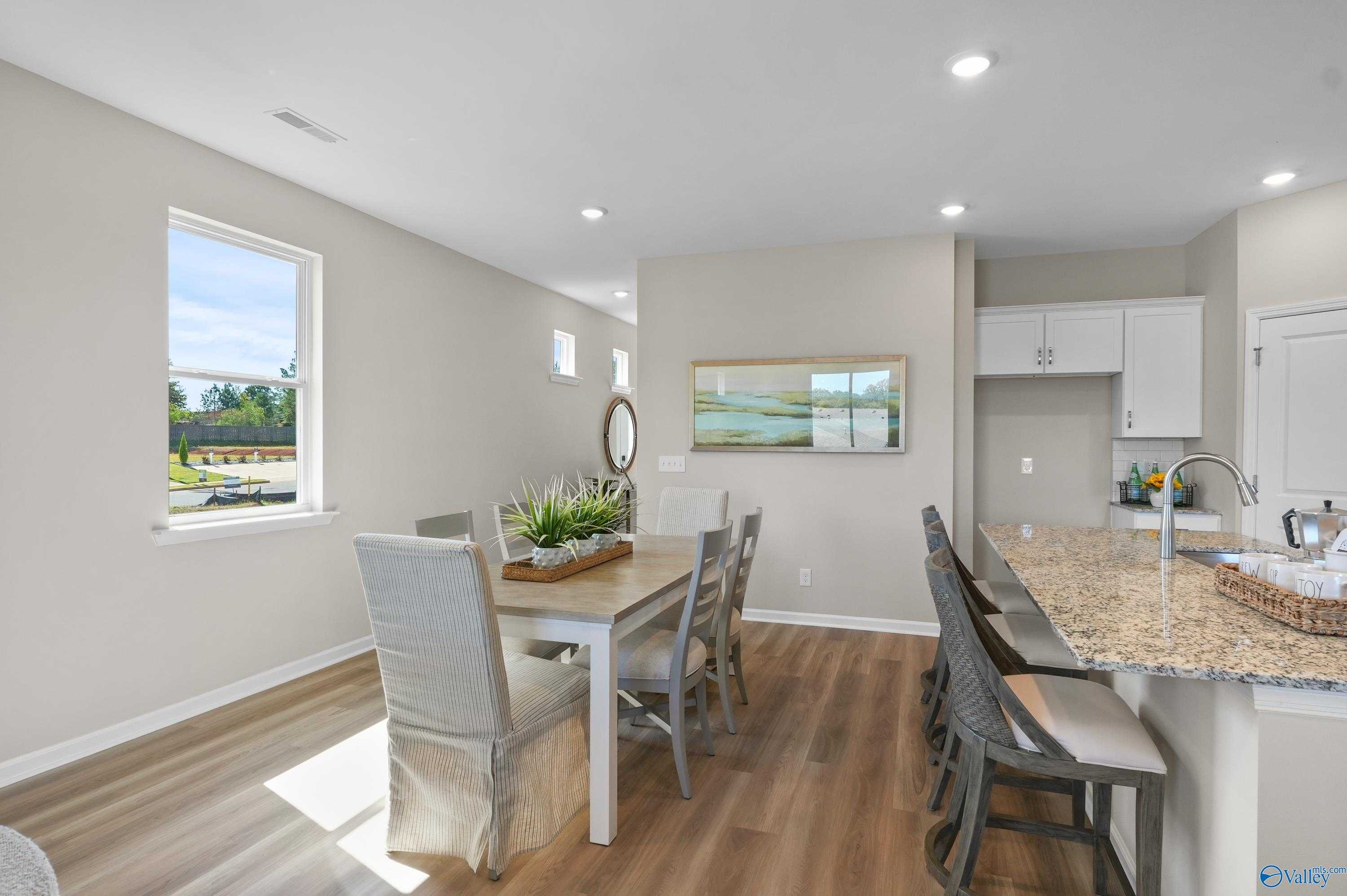Bright dining area with wooden table, upholstered chairs, and potted plants next to granite kitchen in Evermore Homes The Malibu, Madison, Alabama