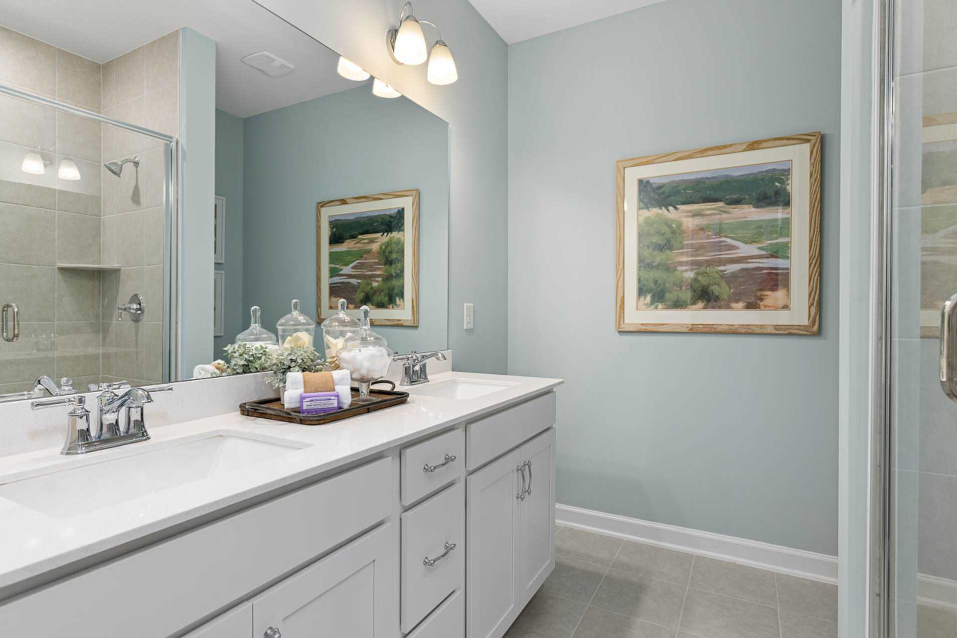 Spacious primary bathroom in Weatherford East, Angier NC by Davidson Homes with light blue walls, double vanity, framed landscape art, and glass shower