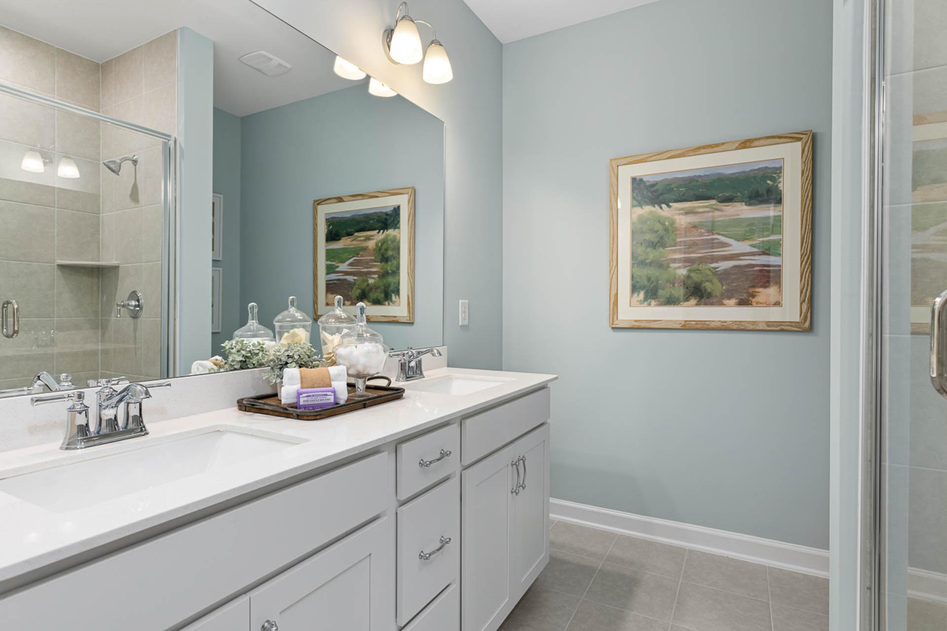 Spacious primary bathroom in Weatherford East, Angier NC by Davidson Homes with light blue walls, double vanity, framed landscape art, and glass shower