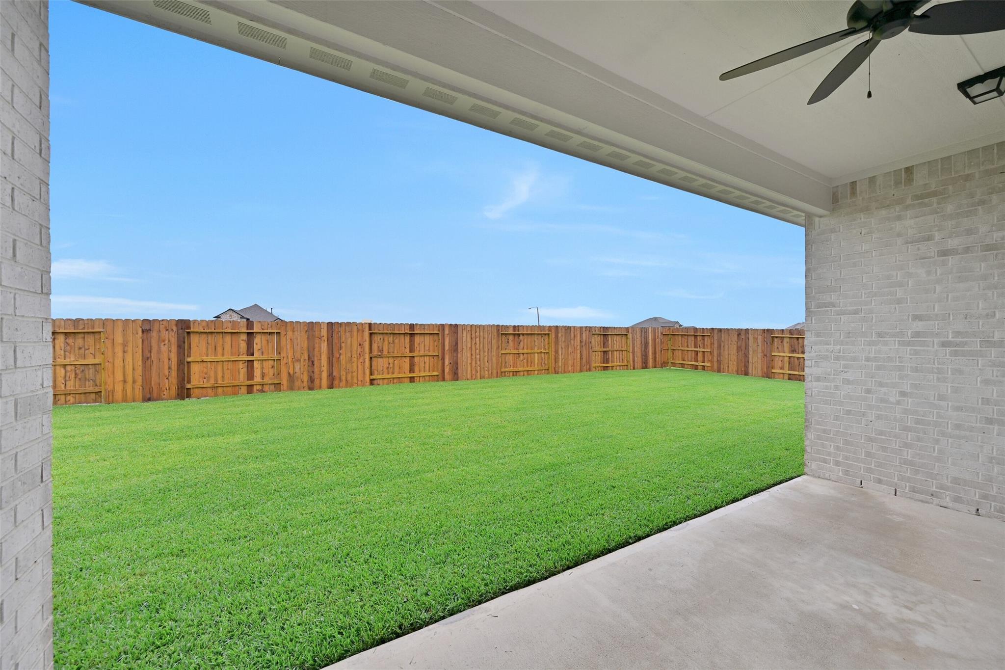 Covered back patio with ceiling fan and brick walls overlooking lush green lawn, wooden fence, and blue sky in Davidson Homes Edward C, Lago Mar, Texas City