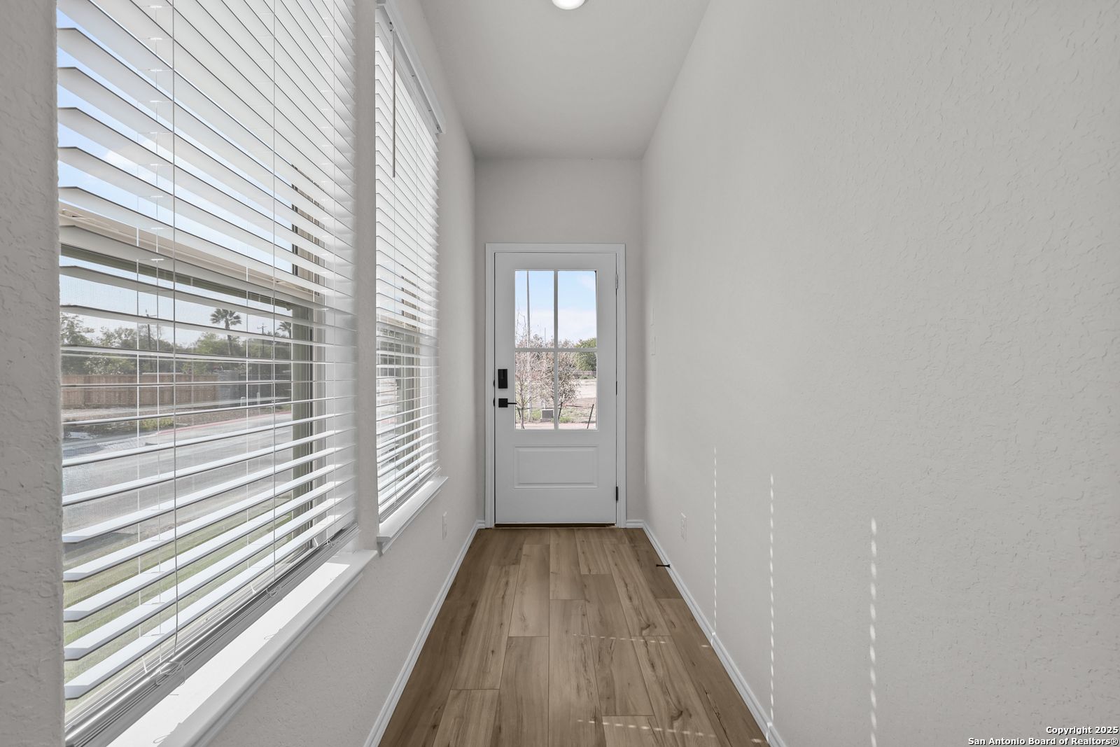 Bright hallway with light wood floors, large windows with white blinds, and glass-paneled white door in Davidson Homes The Florence C, San Antonio