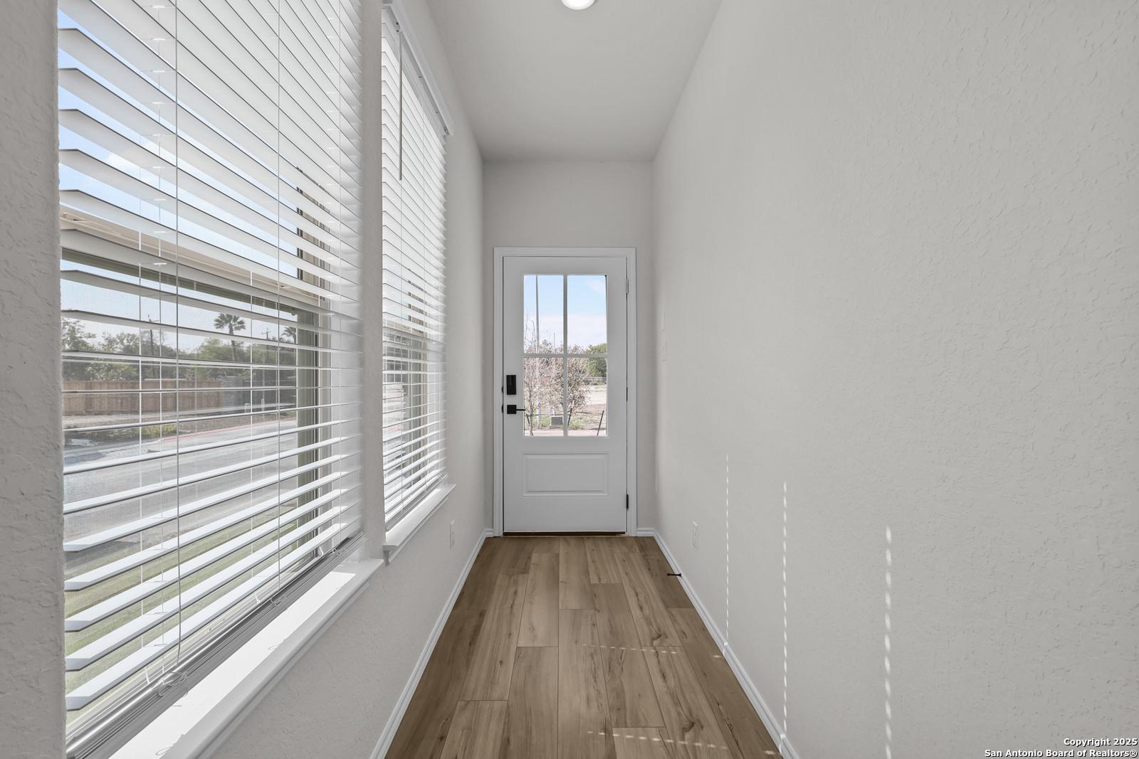 Bright hallway with light wood floors, large windows with white blinds, and glass-paneled white door in Davidson Homes The Florence C, San Antonio