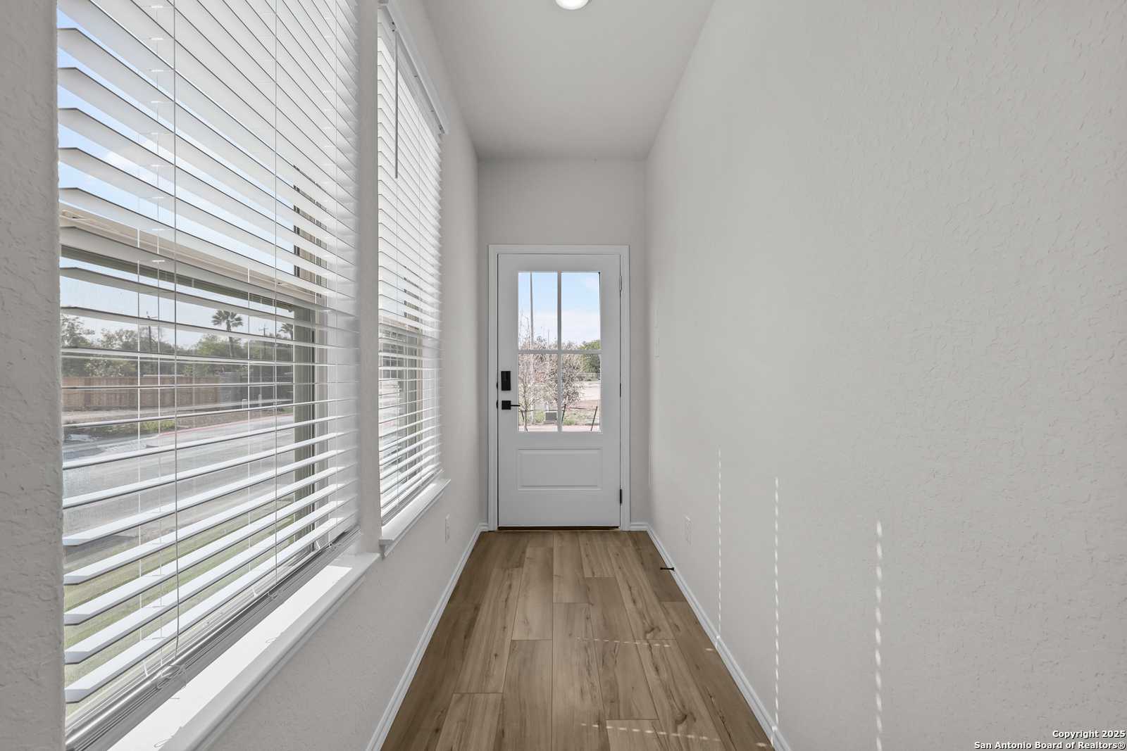 Bright hallway with light wood floors, large windows with white blinds, and glass-paneled white door in Davidson Homes The Florence C, San Antonio