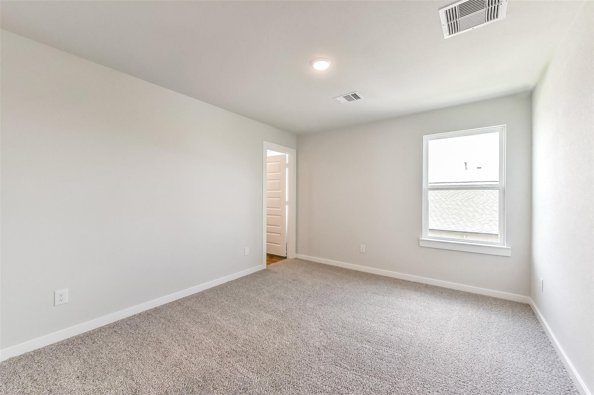 Spacious empty bedroom with light gray walls, beige carpet, large window, and open bath door in Davidson Homes The Tierra C, Dayton Texas