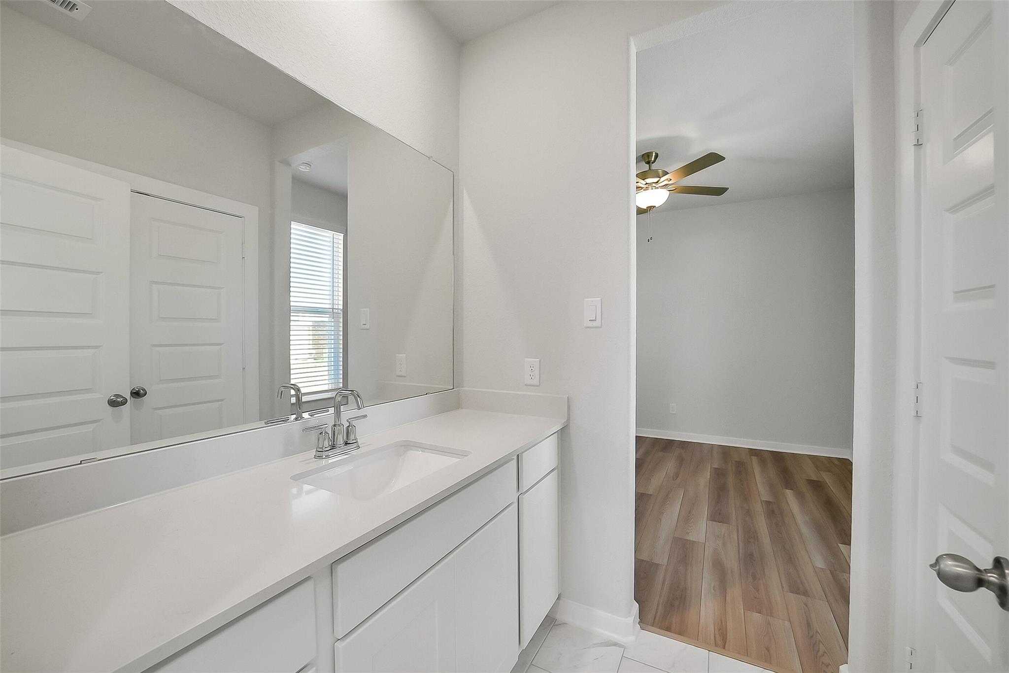 Modern master bathroom with white quartz vanity, single sink, large mirror, and adjacent bedroom in Davidson Homes The Acadia A, Katy TX