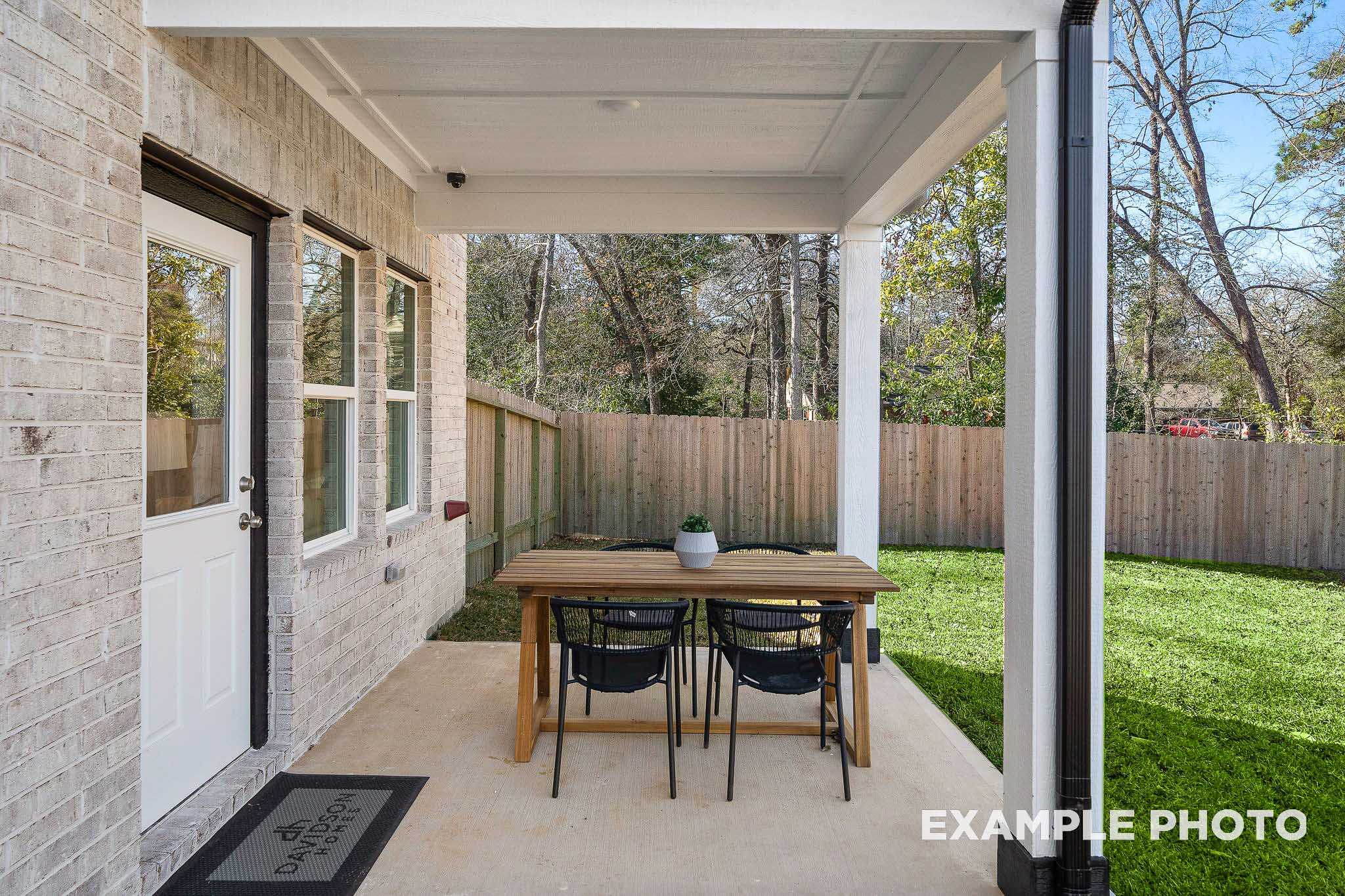 Covered patio of San Marcos Davidson Homes with wooden dining table, chairs, potted plant, white brick exterior, and lush backyard