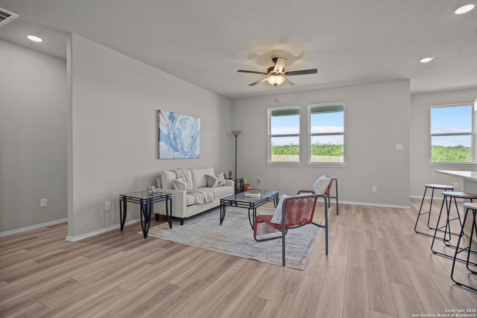 Cozy living room with beige sofa, red accent chair, ceiling fan, and large windows on hardwood floors in Davidson Homes The Asheville H, Converse TX