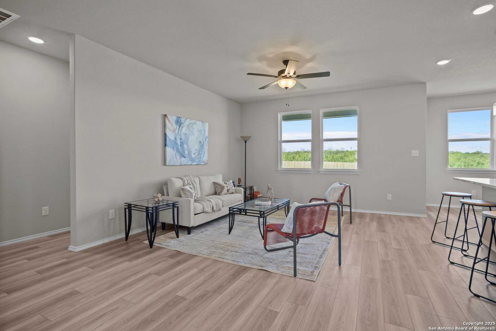 Cozy living room with beige sofa, red accent chair, ceiling fan, and large windows on hardwood floors in Davidson Homes The Asheville H, Converse TX
