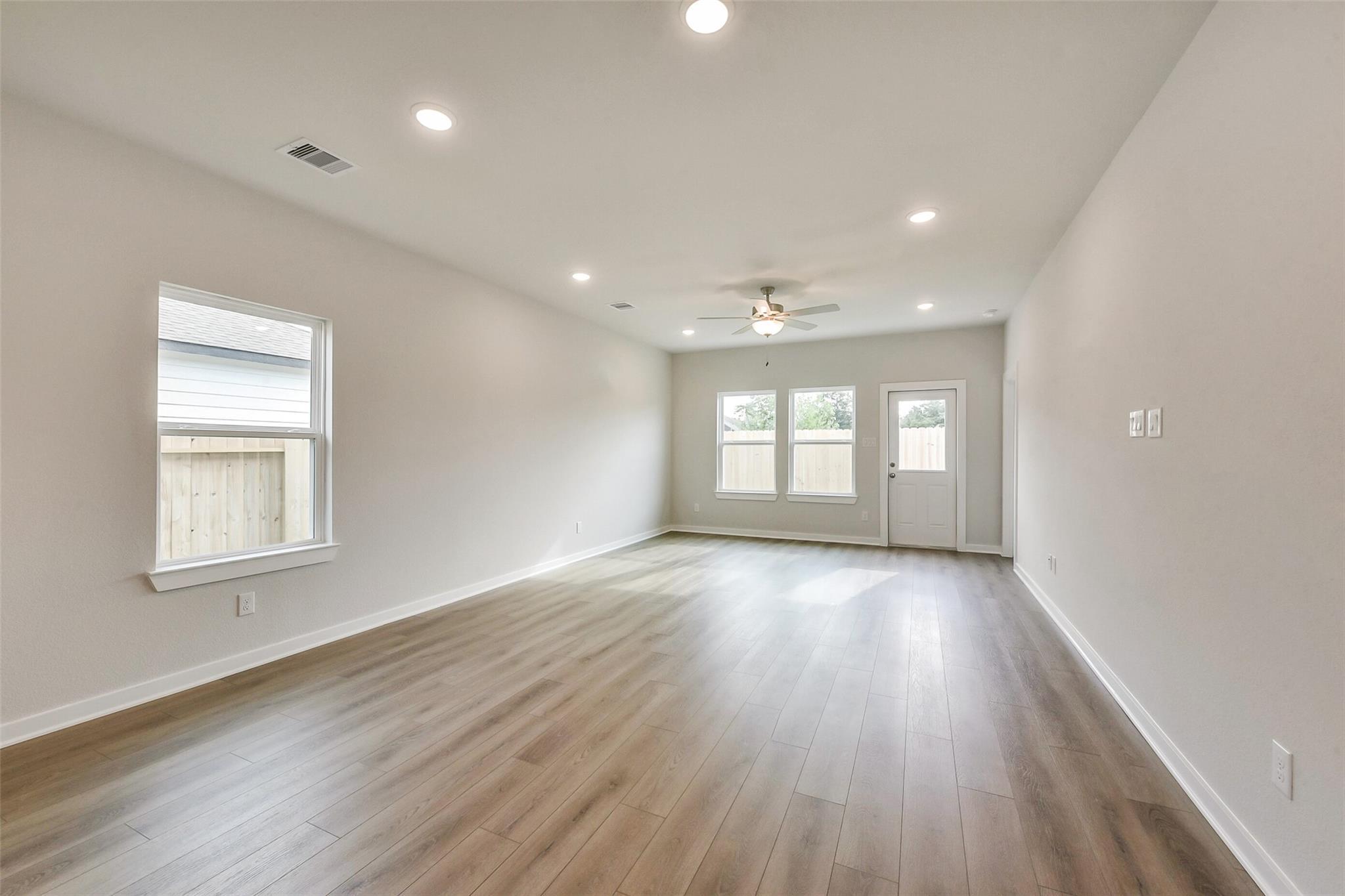 Spacious empty living room with wood laminate floors, ceiling fan, large windows, and patio doors in Davidson Homes The Frio G, Dayton, Texas