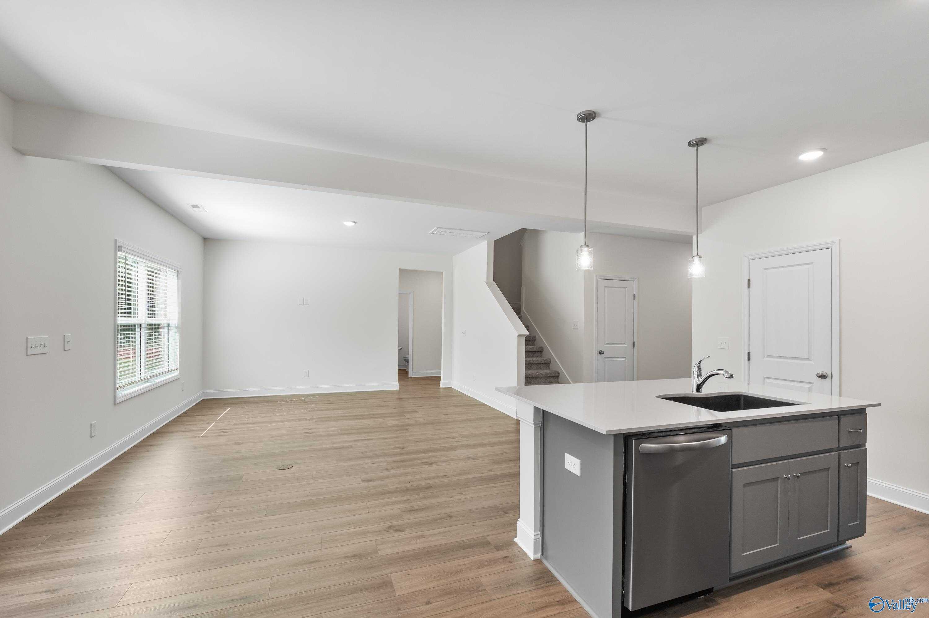 Modern open kitchen with white island, sink, dishwasher, hardwood floors, and staircase in The Shelby A home, Athens, Alabama