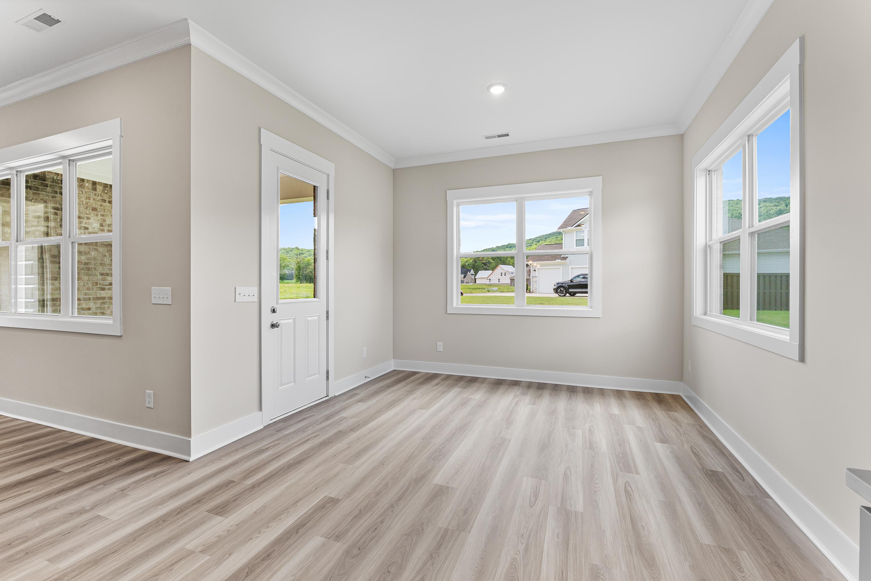 Bright sunlit room in The Oxford A home featuring beige walls, large windows with green views, white door, and light wood floors