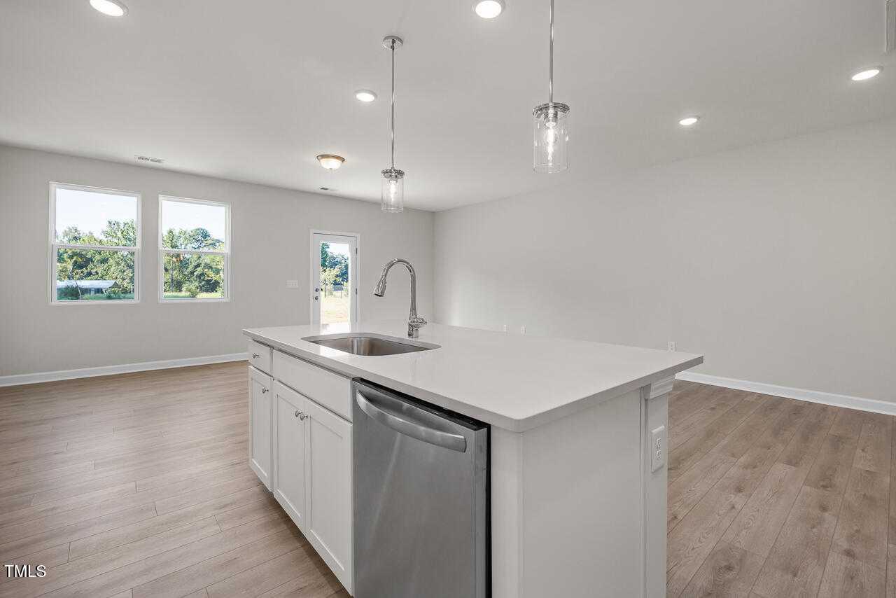 Modern white kitchen island with quartz countertop, stainless dishwasher, and hardwood floors in The Warren Interior by Davidson Homes, Fuquay-Varina, NC