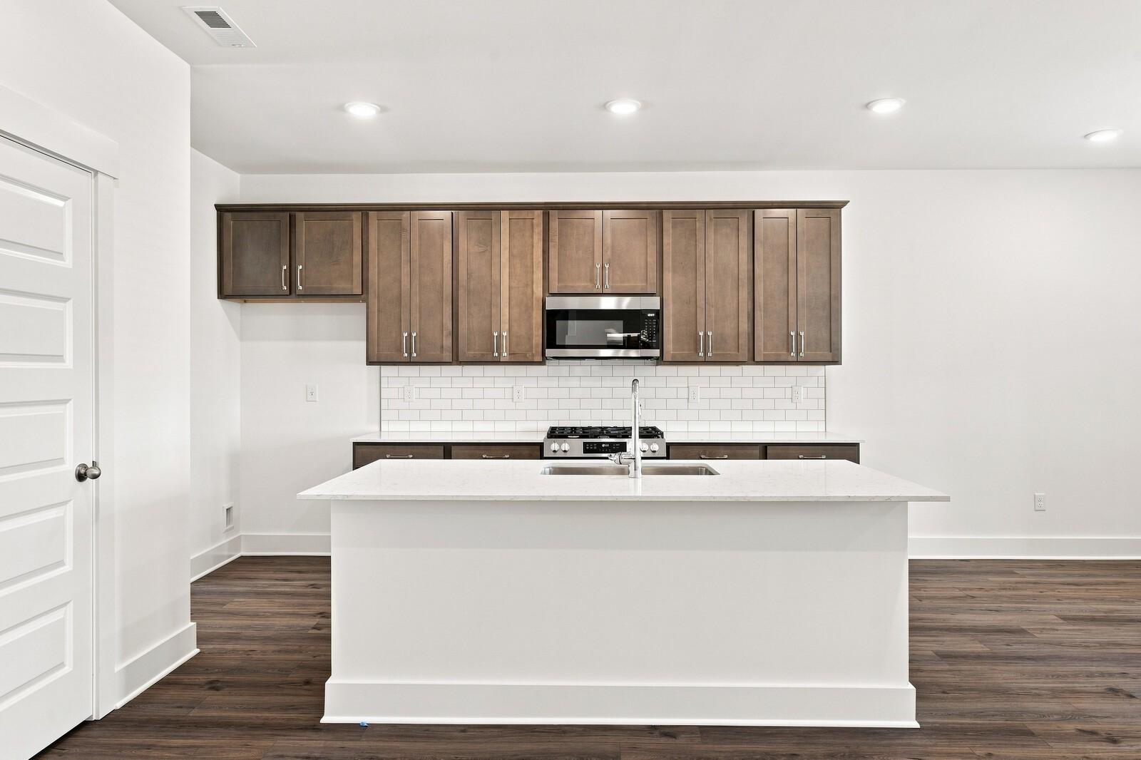 White kitchen island with sink, dark wood cabinets, subway tile backsplash, and stainless appliances in Davidson Homes Gallatin home