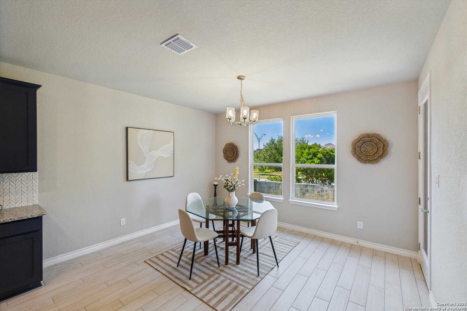 Bright dining room with round glass table, four white chairs, chandelier, and garden-view windows in The Jennings H, Ladera, San Antonio