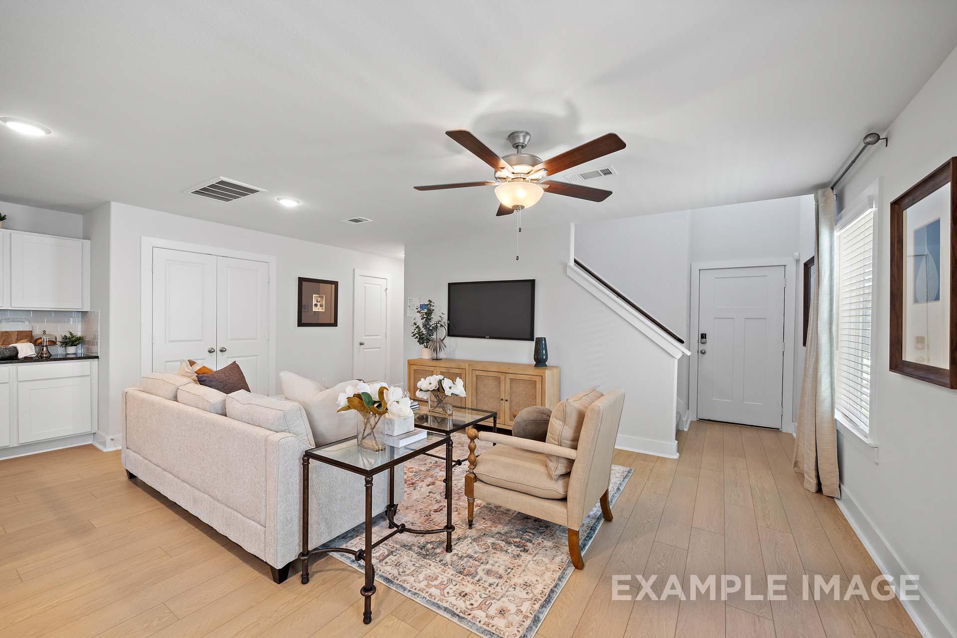 Spacious living room in The Rose B by Davidson Homes with beige sofa, armchair, ceiling fan, wooden floors, and adjacent white kitchen