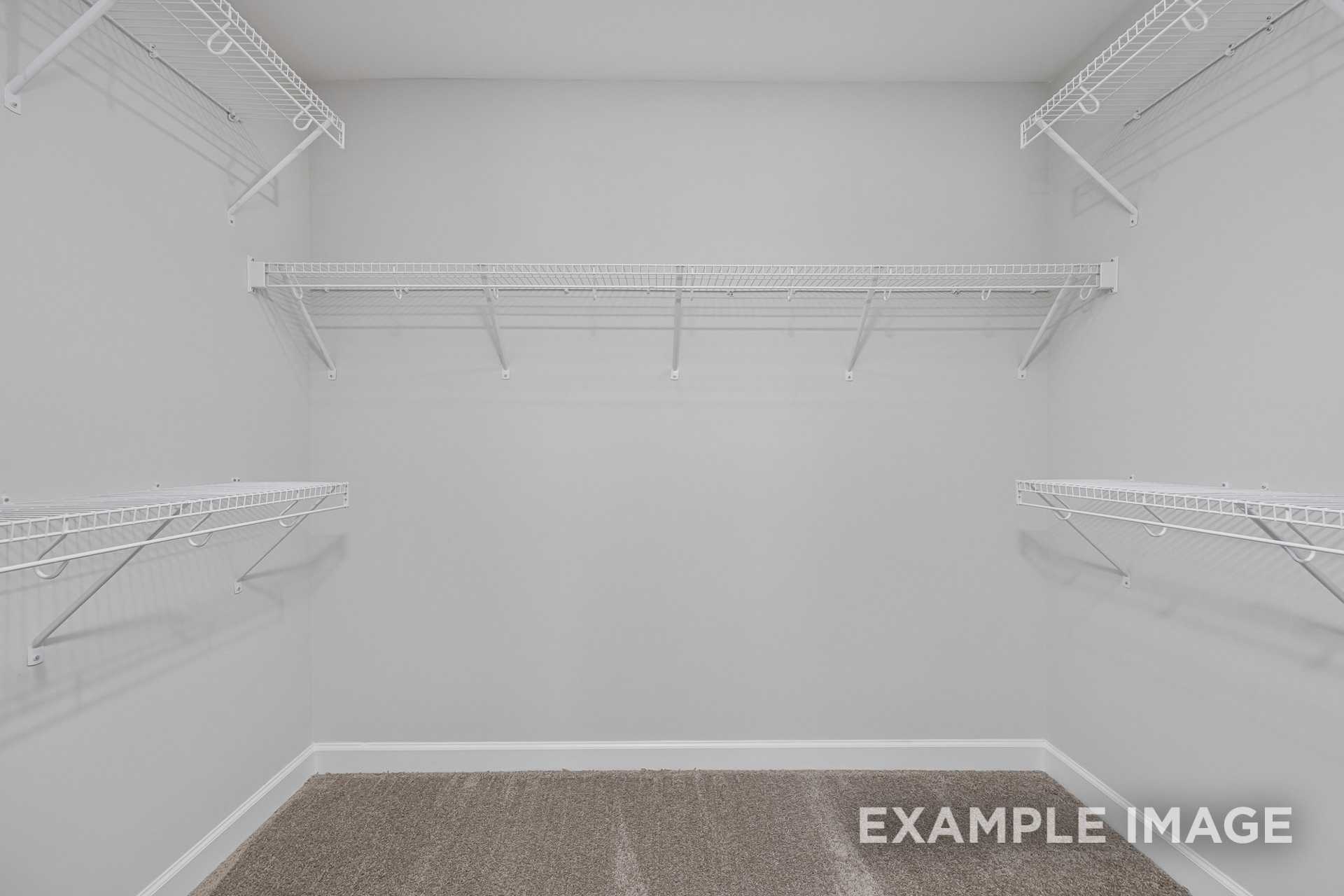 Spacious walk-in closet in The Adalynn A master suite featuring white wire shelving, hanging rods on light gray walls, brown carpet floor