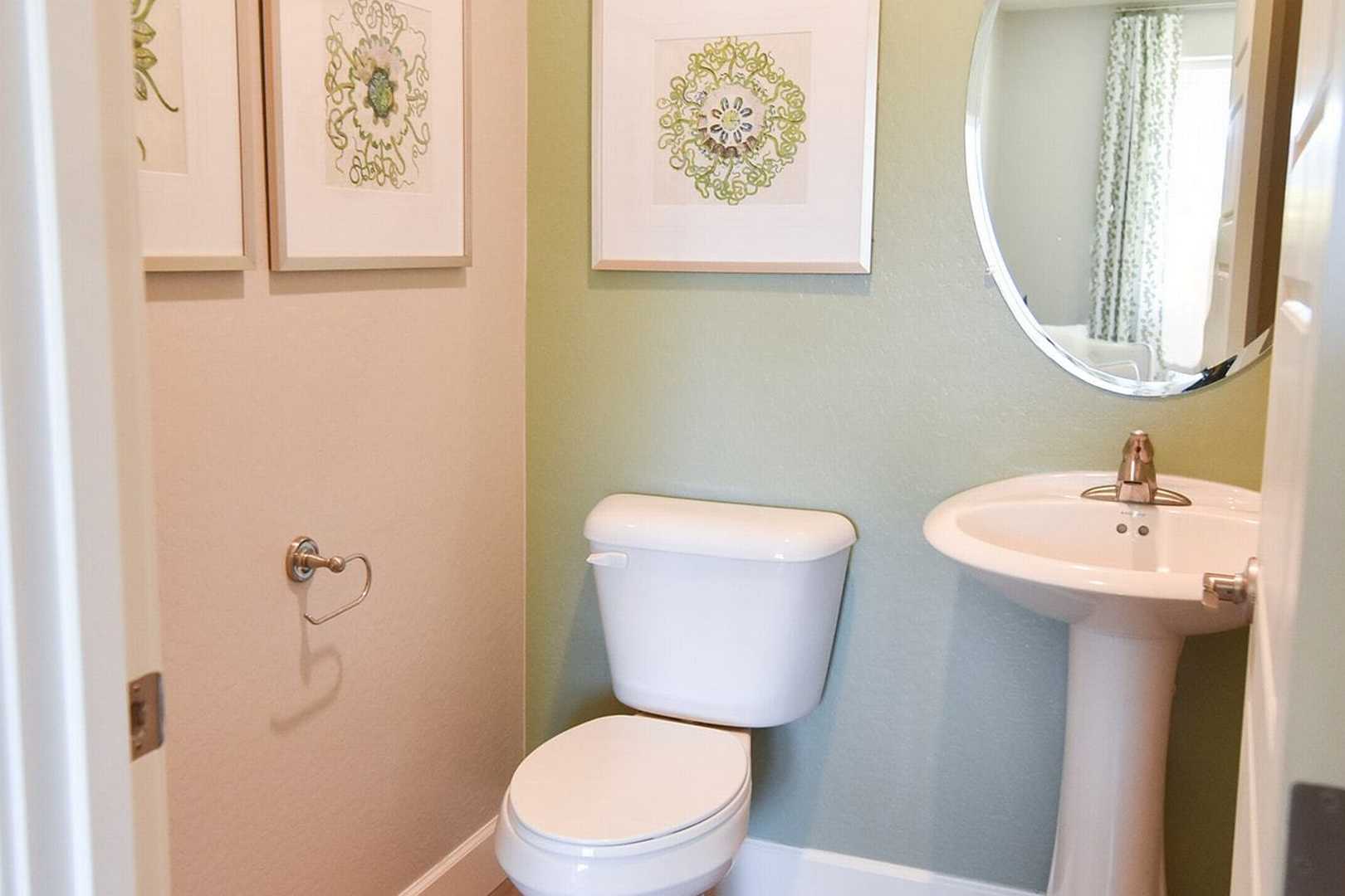 Cozy powder room in The Summit B with pedestal sink, white toilet, round mirror, and green floral wall art