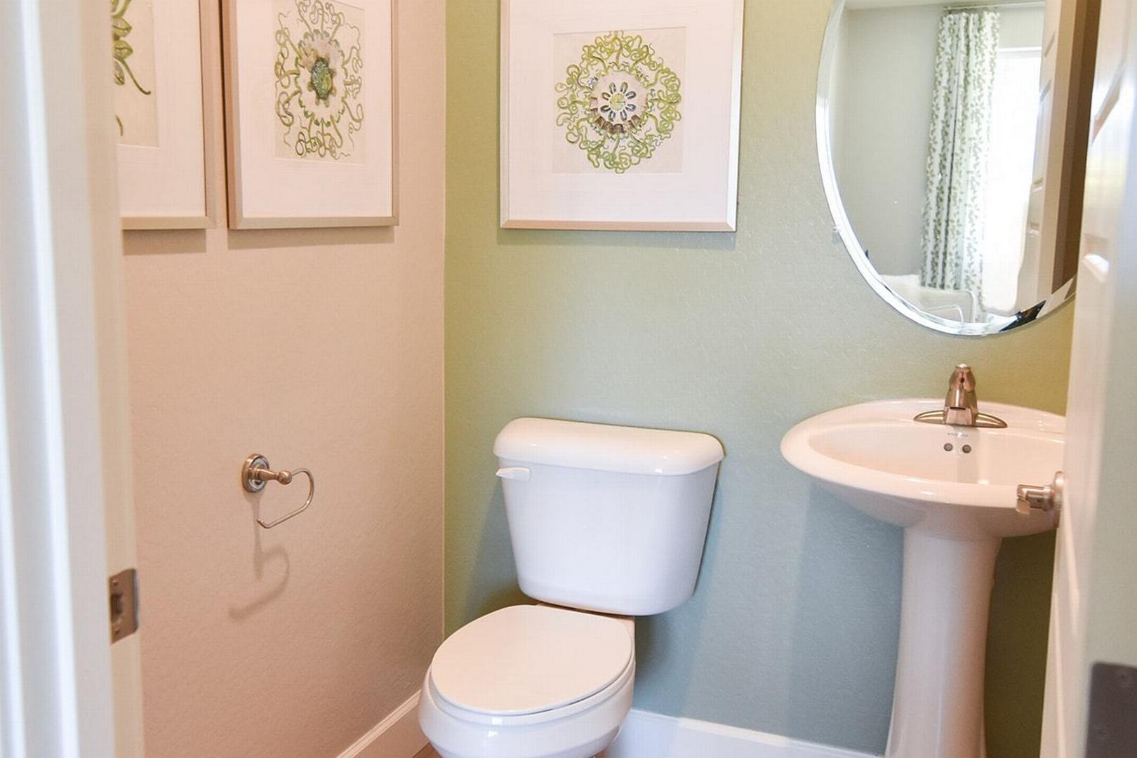 Compact powder room in The Summit E home featuring pedestal sink, white toilet, round mirror, and green floral wall art