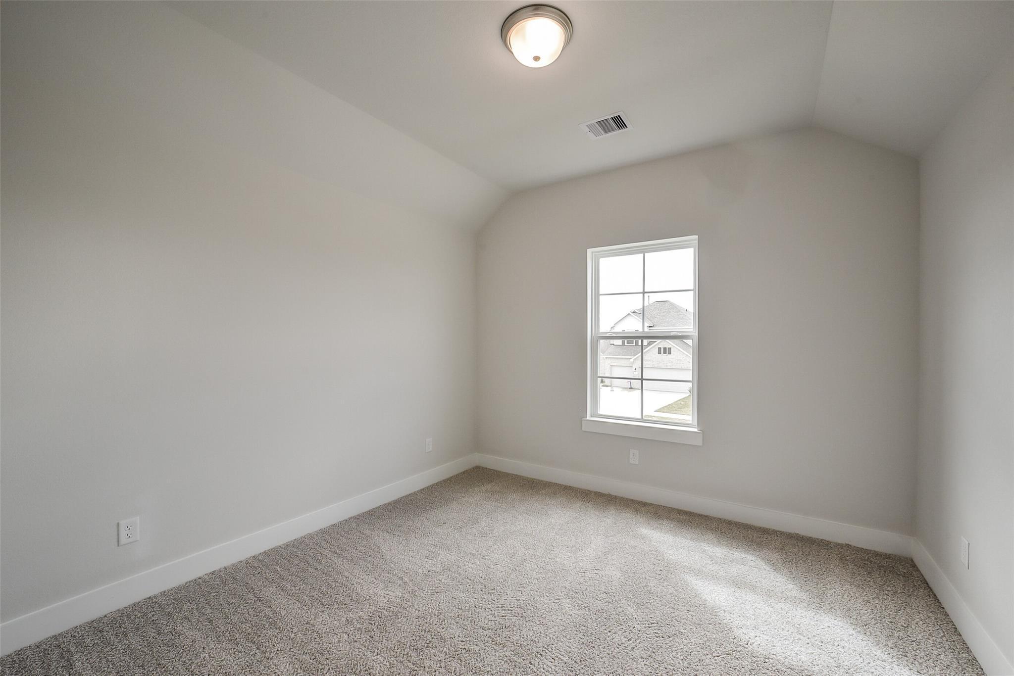 Bright empty bedroom with neutral walls, beige carpet, sloped ceiling, and window view in Davidson Homes The Philip B, Rosharon, Texas