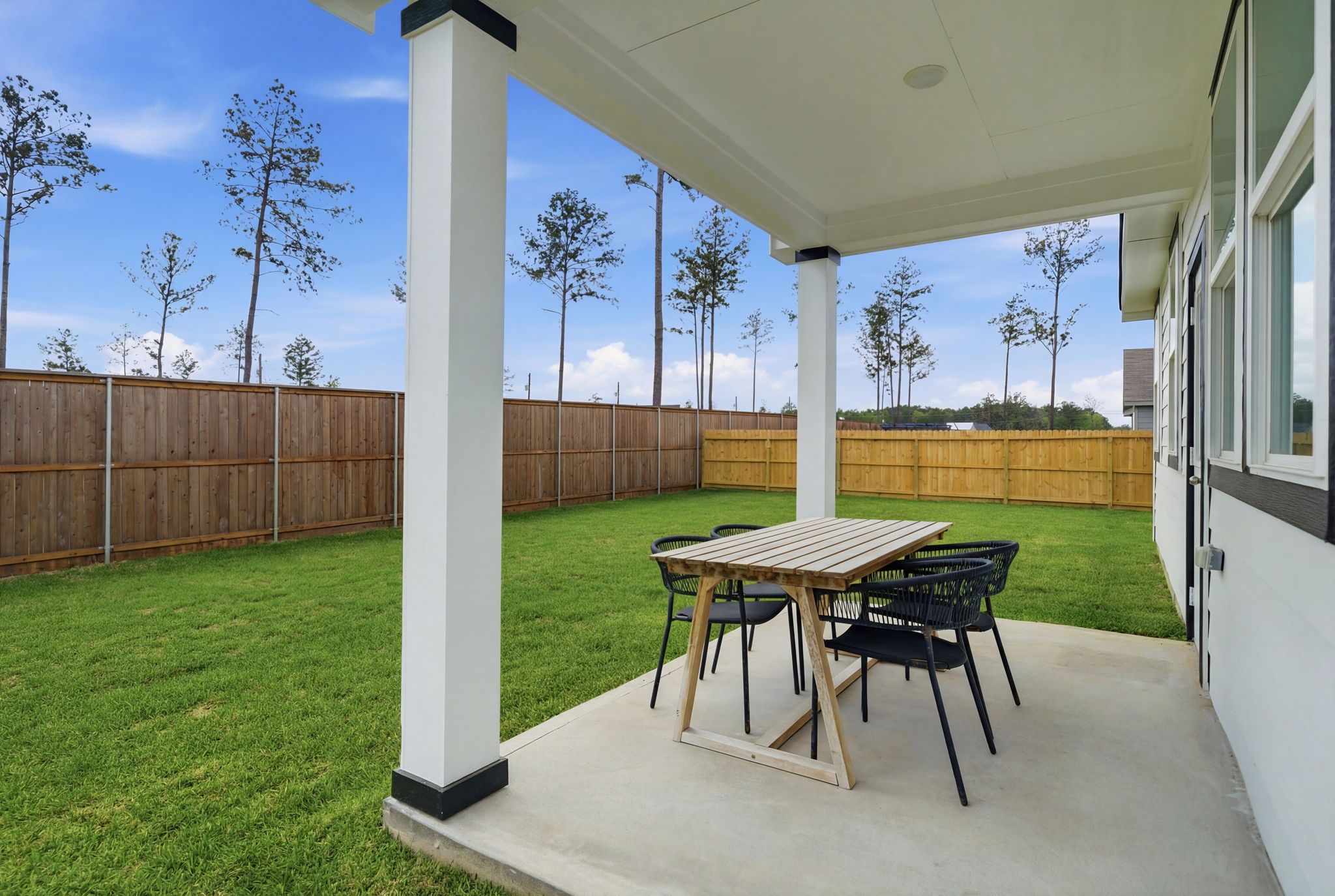 Covered backyard patio with wooden dining table, rattan chairs, lush green lawn, and pine trees at Spring Branch Crossing in Conroe Texas