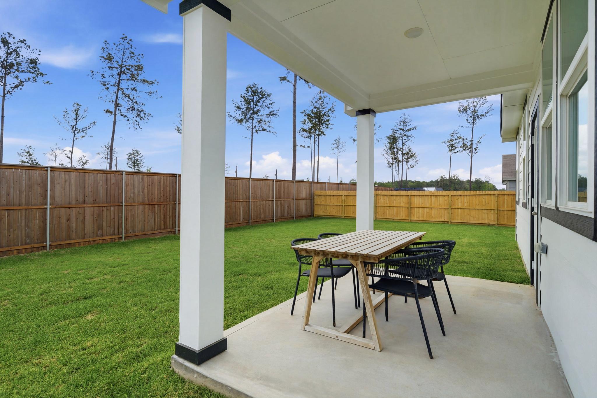 Covered backyard patio with wooden dining table, rattan chairs, lush green lawn, and pine trees at Spring Branch Crossing in Conroe Texas