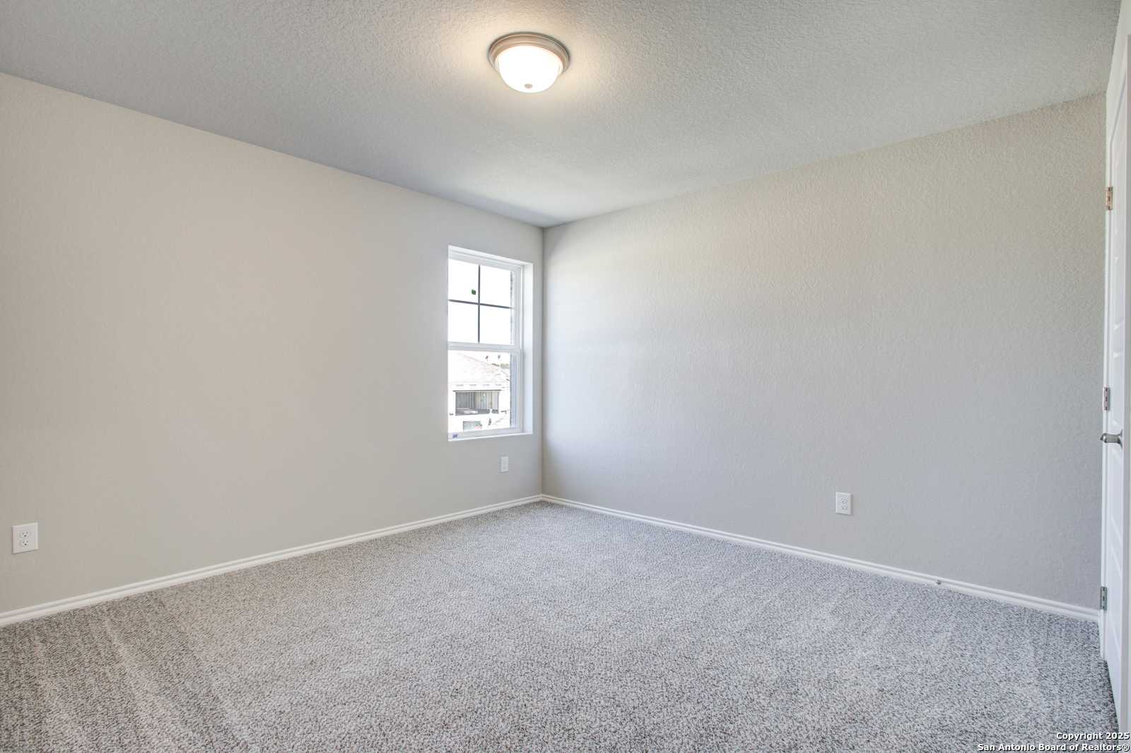 Spacious empty bedroom with beige walls, gray carpet, large window, and ceiling light in Davidson Homes The Douglas G, San Antonio