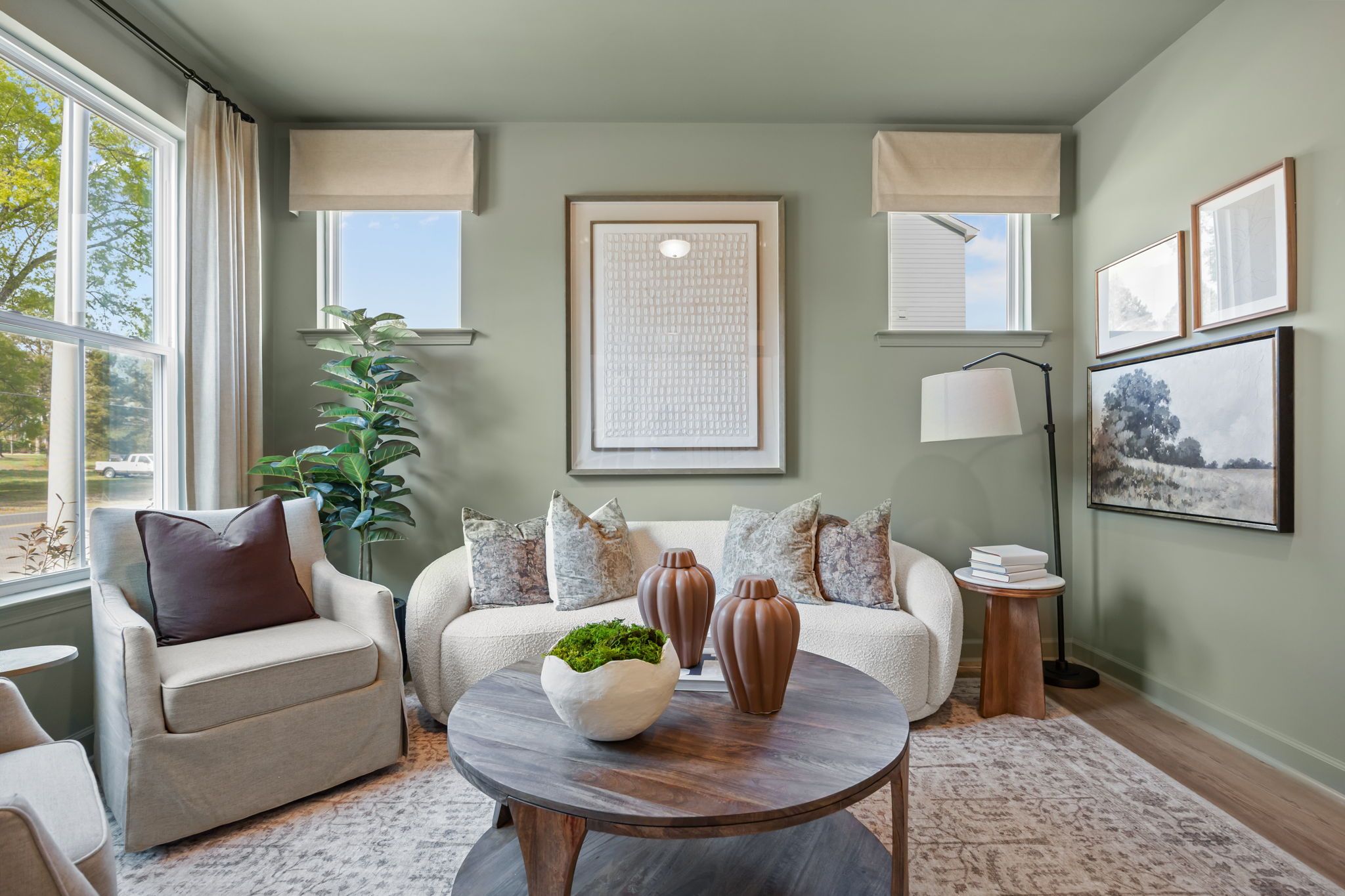 Cozy living room in Bradford Raleigh NC with sage green walls, curved white sofa, round wood coffee table, and large windows by Davidson Homes