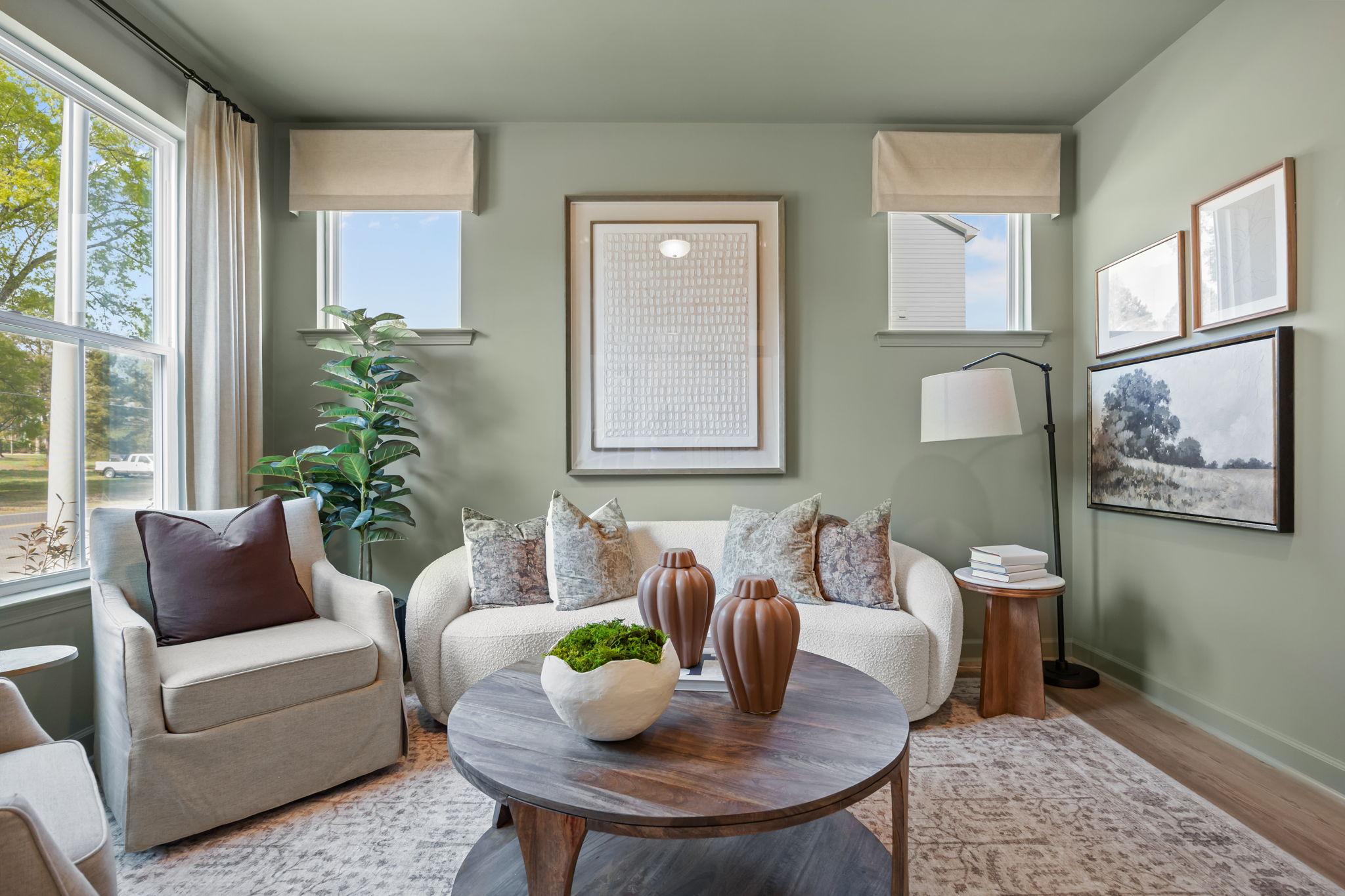 Cozy living room in Bradford Raleigh NC with sage green walls, curved white sofa, round wood coffee table, and large windows by Davidson Homes
