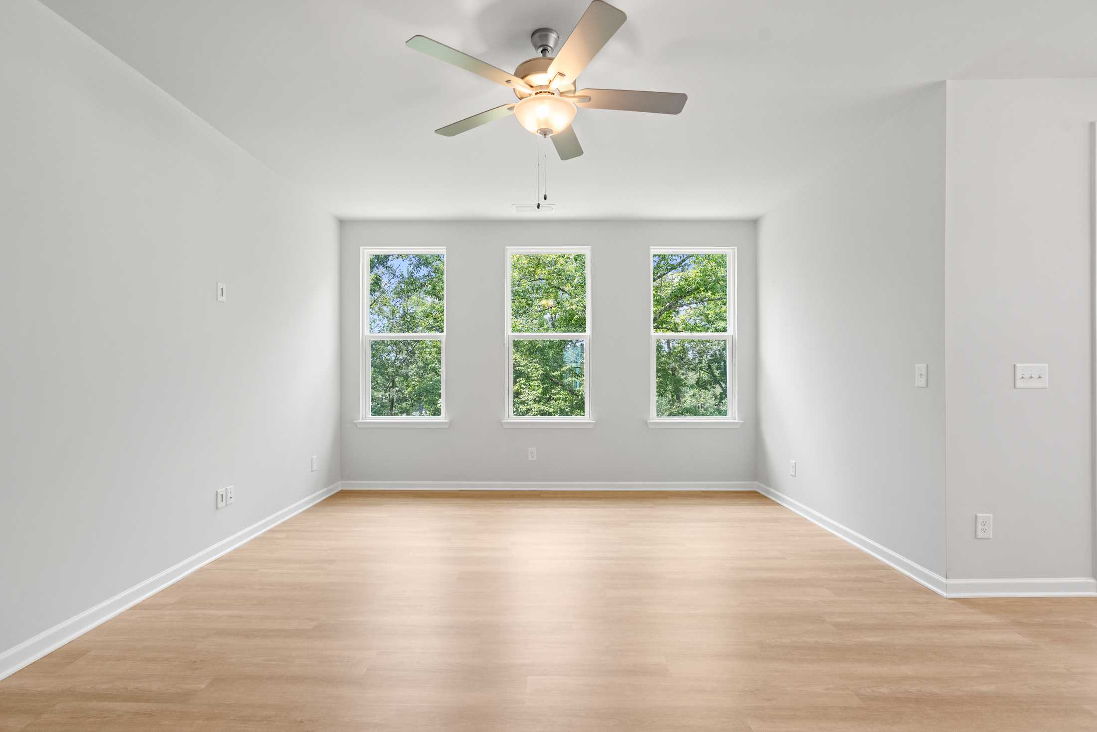 Spacious empty bedroom in The Ash B home with hardwood floors, white walls, three large windows overlooking greenery, and centered ceiling fan