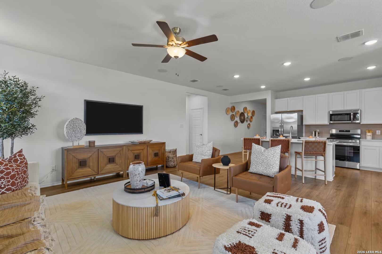 Open-concept living room and kitchen with white cabinets, stainless appliances, leather seating, and ceiling fan in The Daphne K home, San Antonio
