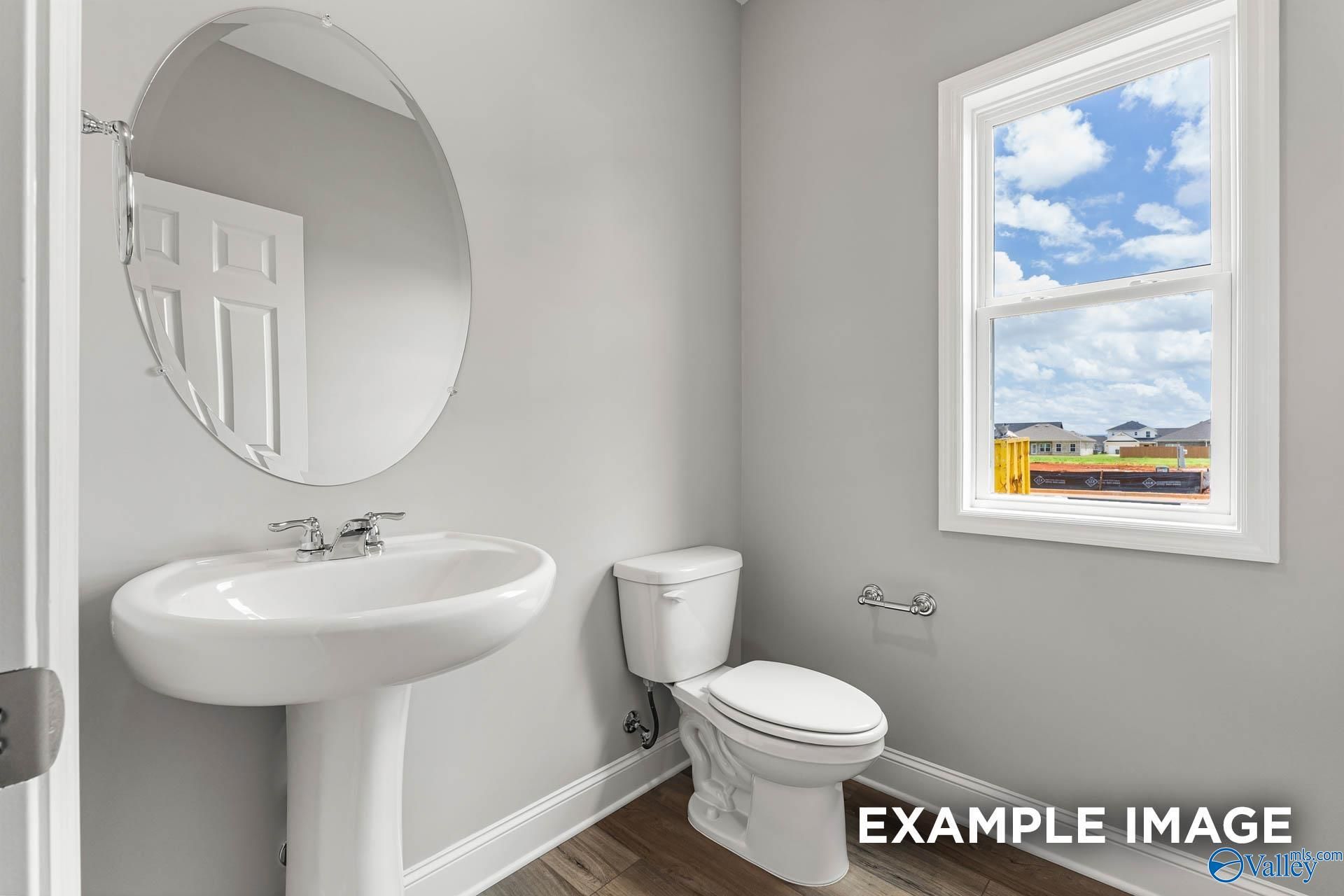 Modern half bath with white pedestal sink, round mirror, toilet, and window view in Davidson Homes The Chelsea A, Athens, Alabama
