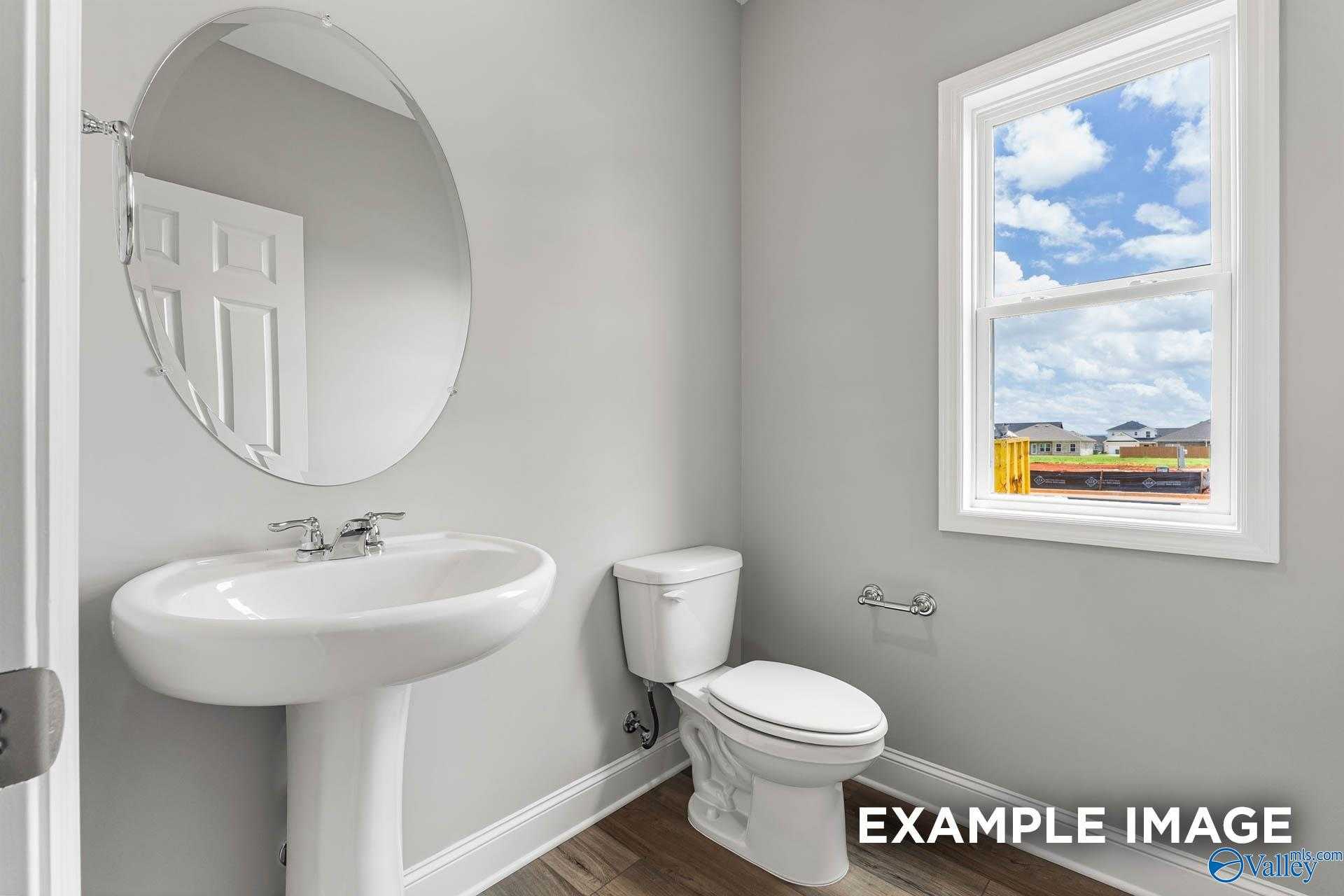 Modern half bath with white pedestal sink, round mirror, toilet, and window view in Davidson Homes The Chelsea A, Athens, Alabama