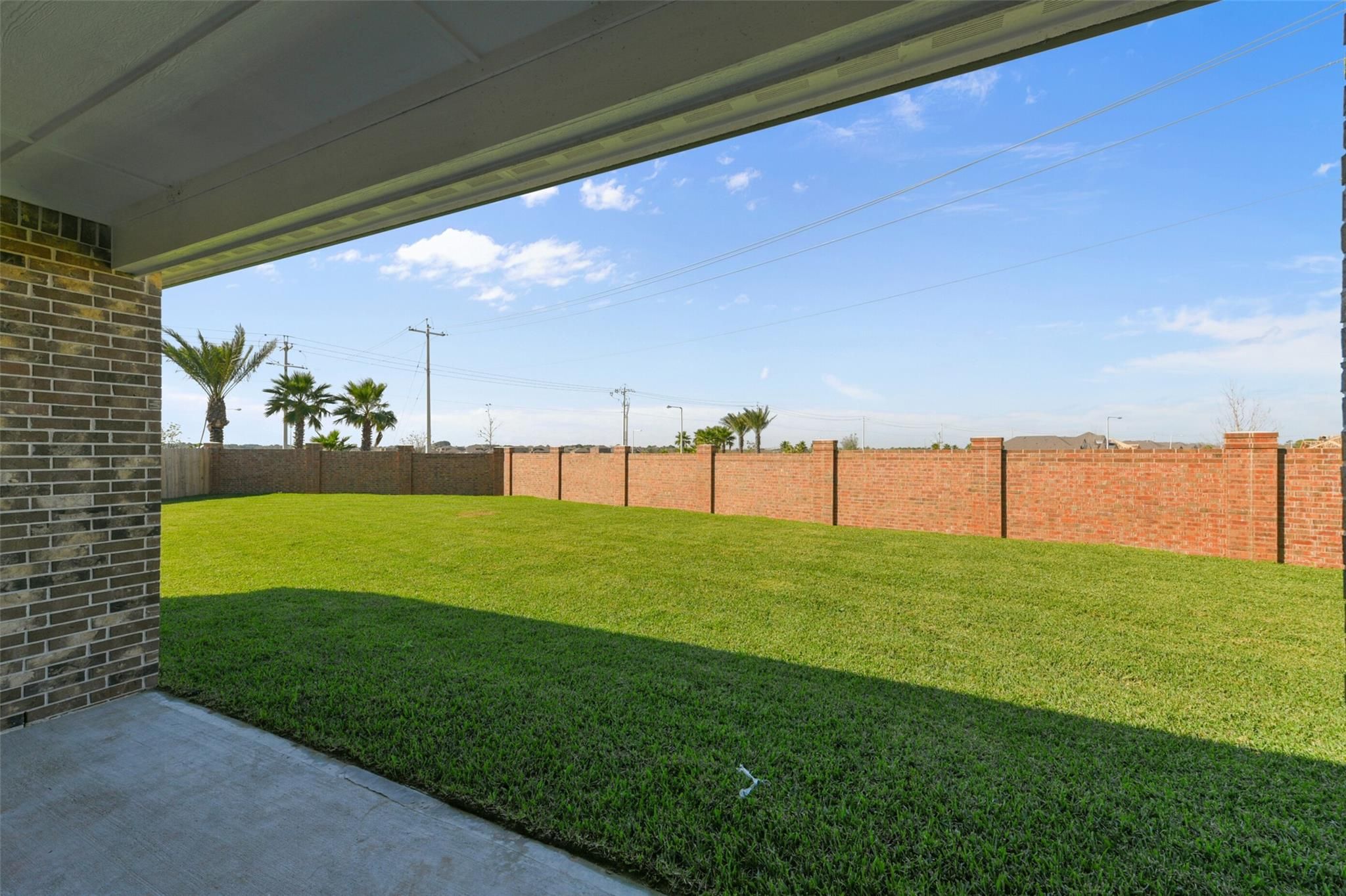 Covered patio with lush green lawn, brick privacy fence, and palm trees in Davidson Homes The Edward C, Lago Mar, Texas