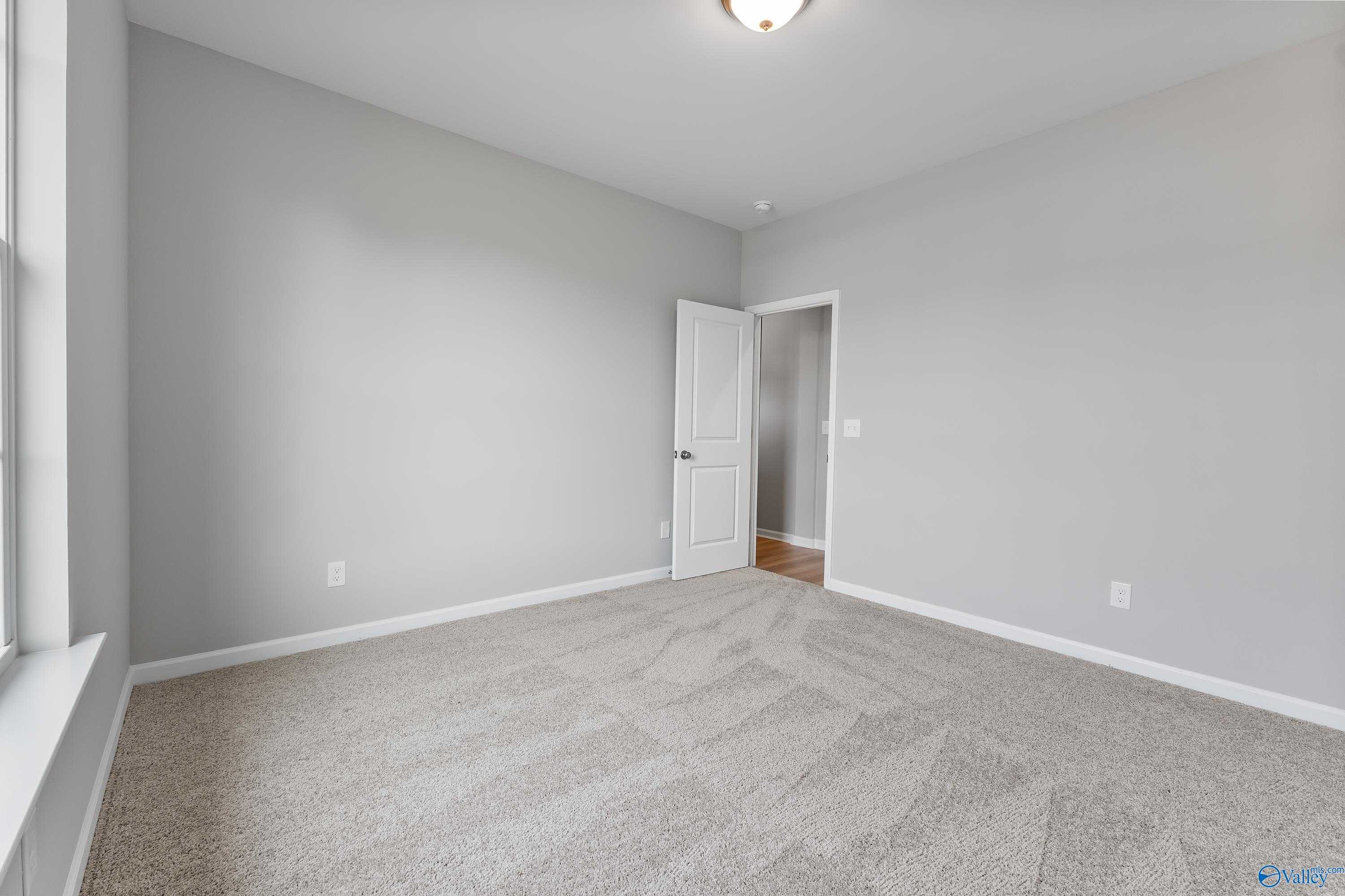 Empty bedroom with light gray walls, beige carpet, adjacent door, and window in Evermore Homes The Grace, Madison Alabama