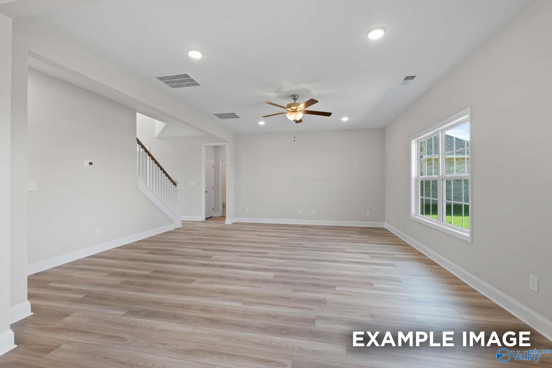 Spacious empty living room with light wood floors, ceiling fan, large windows overlooking lawn, and staircase in The Madison A, Toney, AL