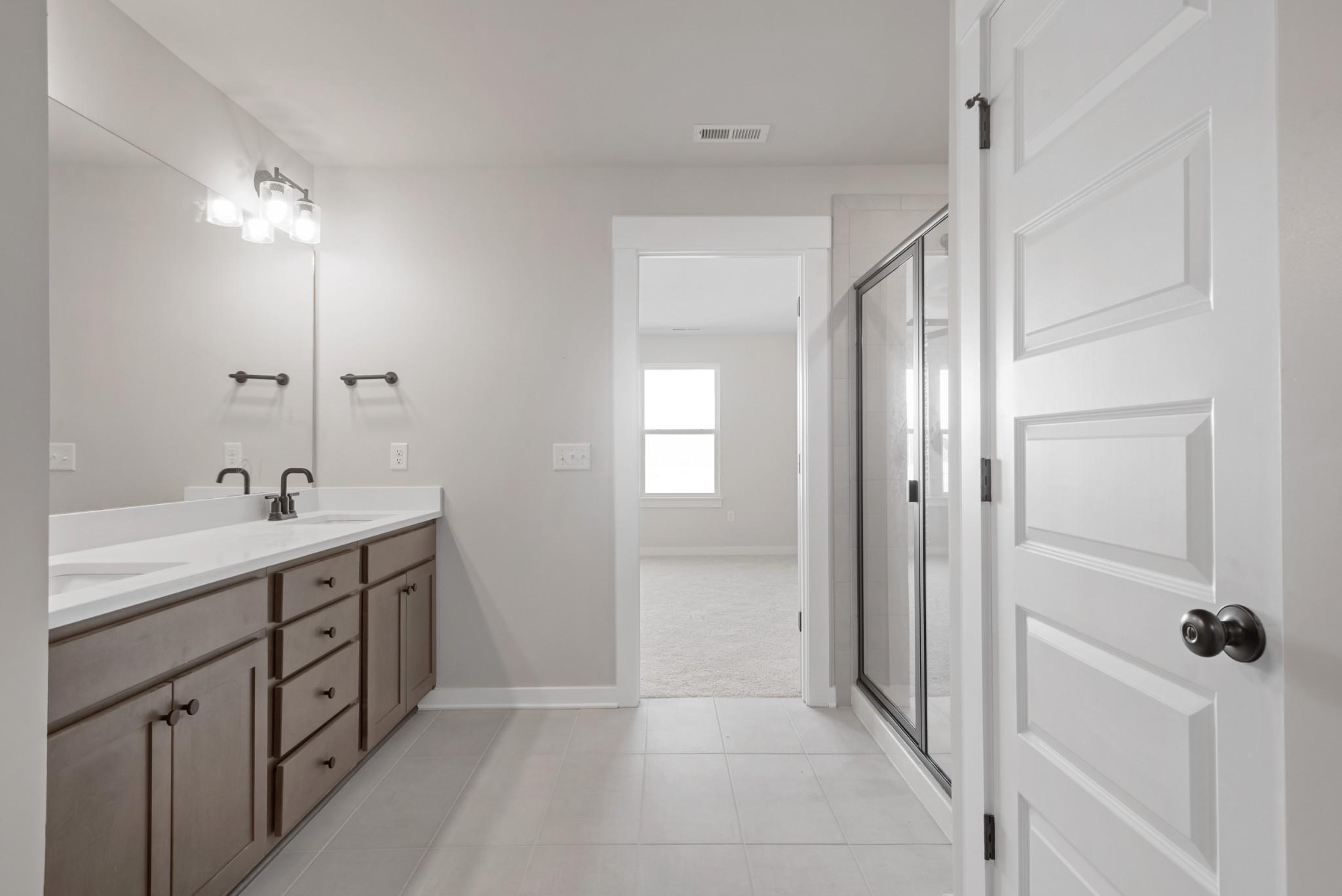Modern master bathroom with double vanity, frameless glass shower, and adjacent bedroom in Davidson Homes The Willow B, Calista Farms