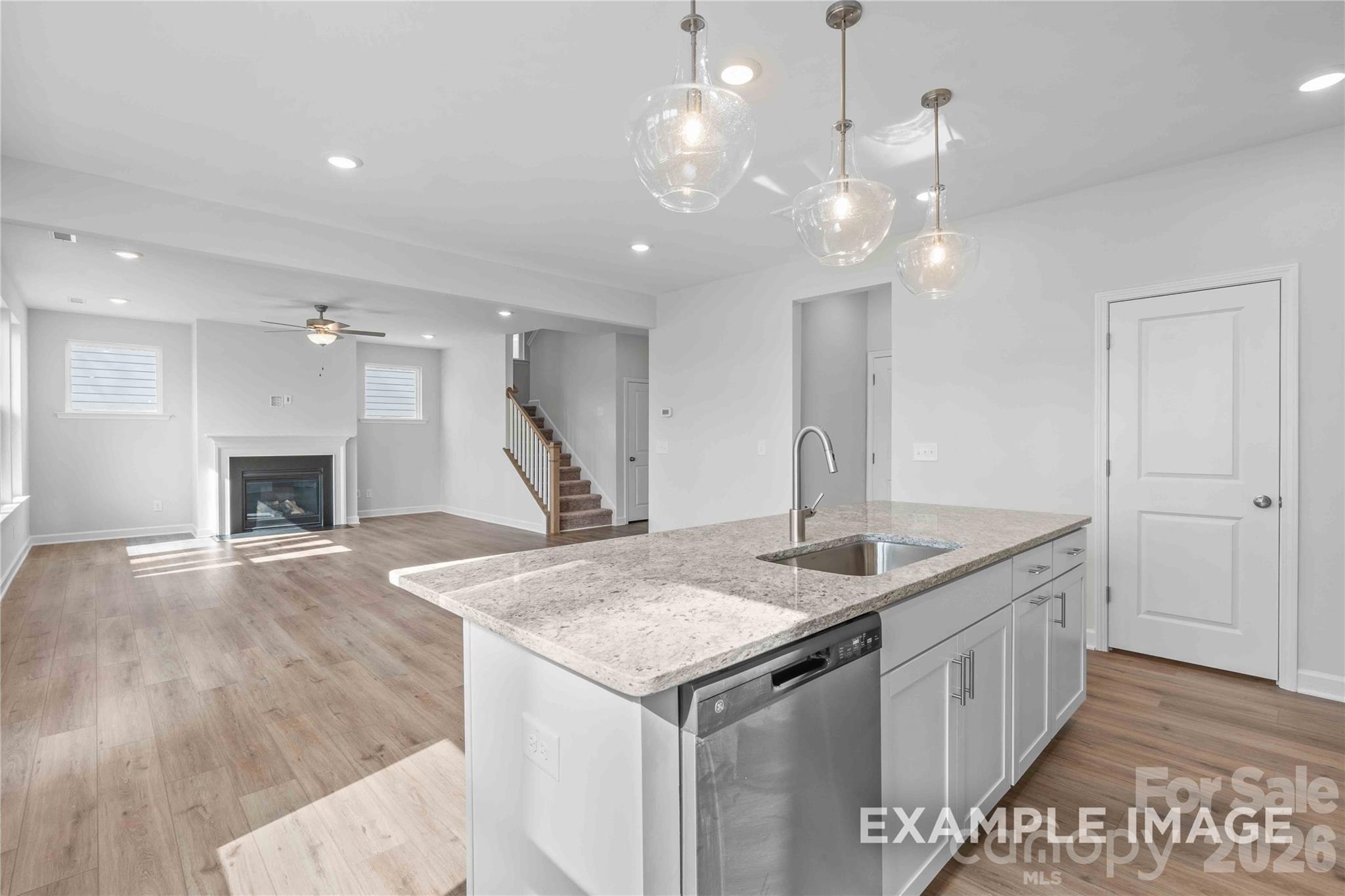 Open-concept kitchen with quartz island, stainless dishwasher, and pendant lights flowing to living room fireplace in The Chestnut D, Belmont, NC