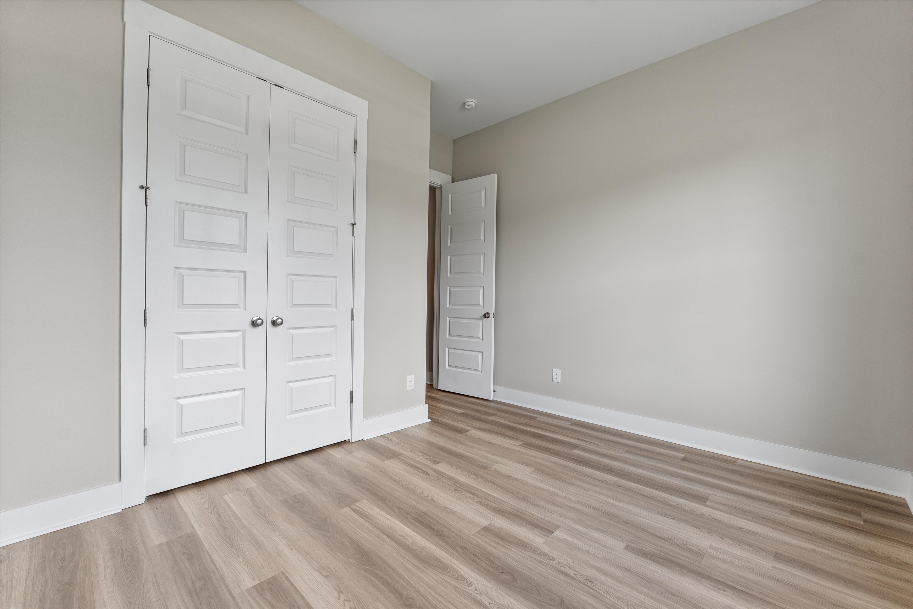 Spacious empty bedroom in The Oxford with white double doors, neutral beige walls, and light wood laminate flooring