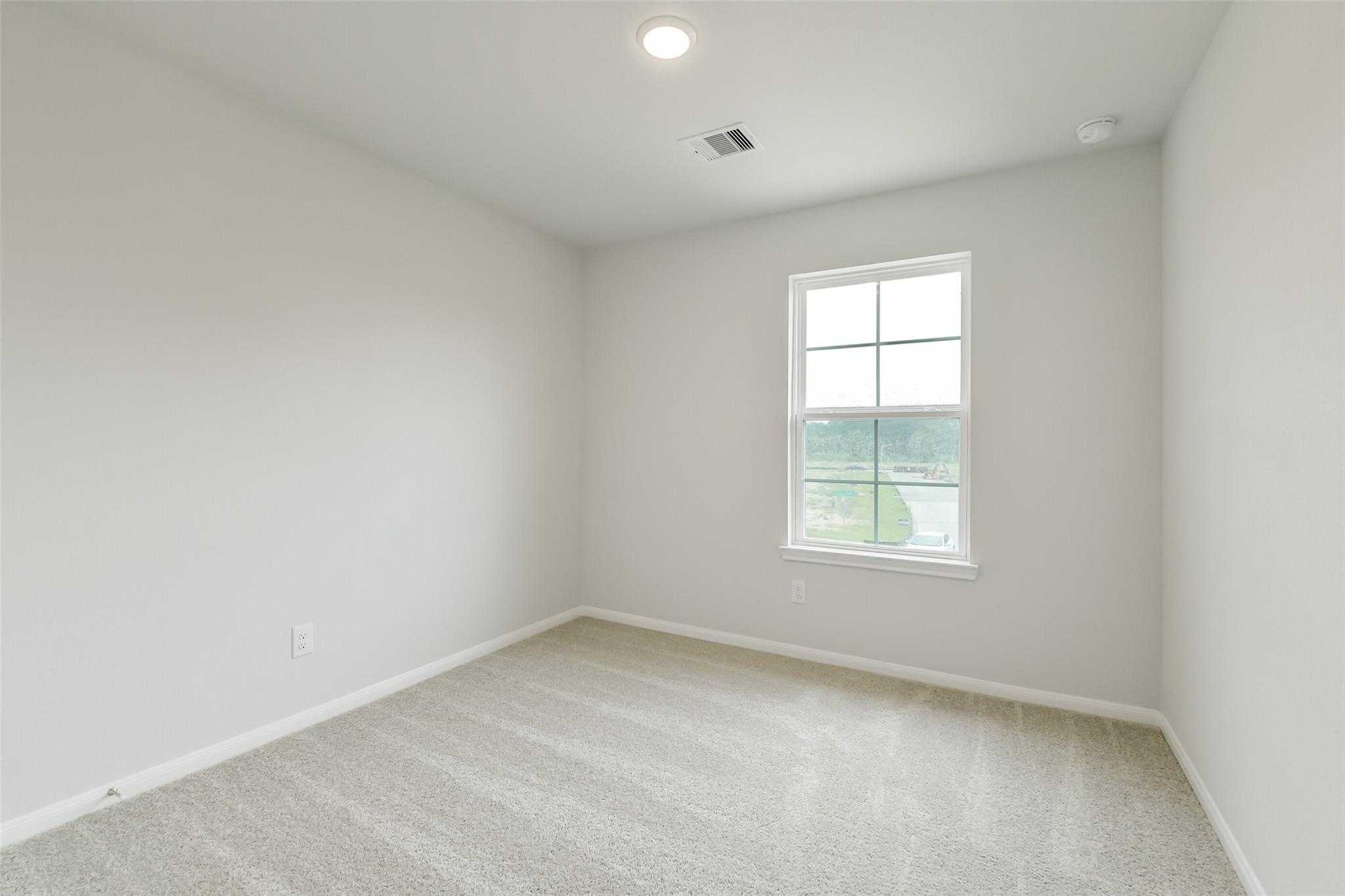 Bright secondary bedroom with neutral walls, beige carpet, and large window overlooking greenery in The Brazos F Conroe TX home