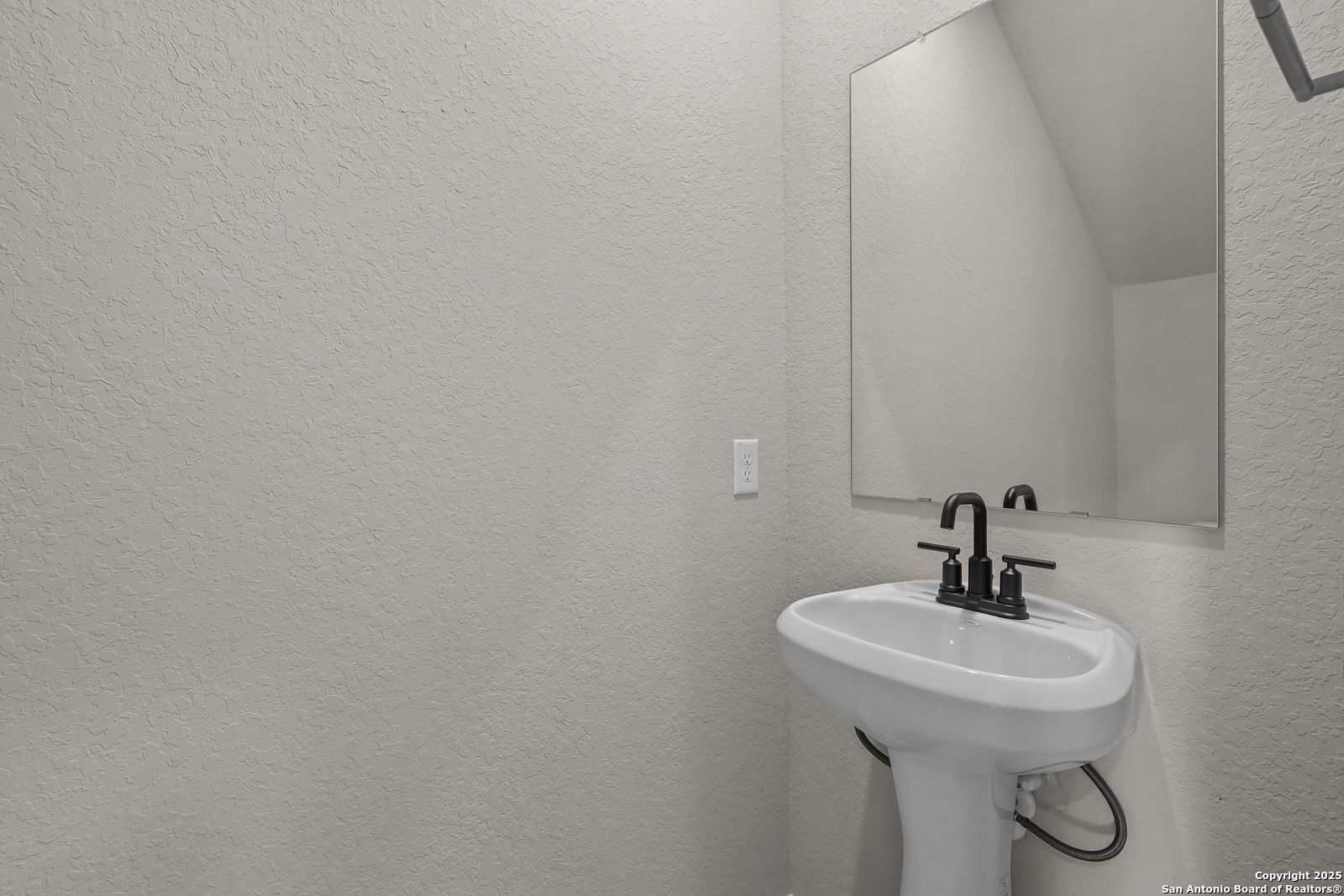 Elegant powder room with white pedestal sink, black faucet, and frameless mirror in Davidson Homes The Brazos C, San Antonio