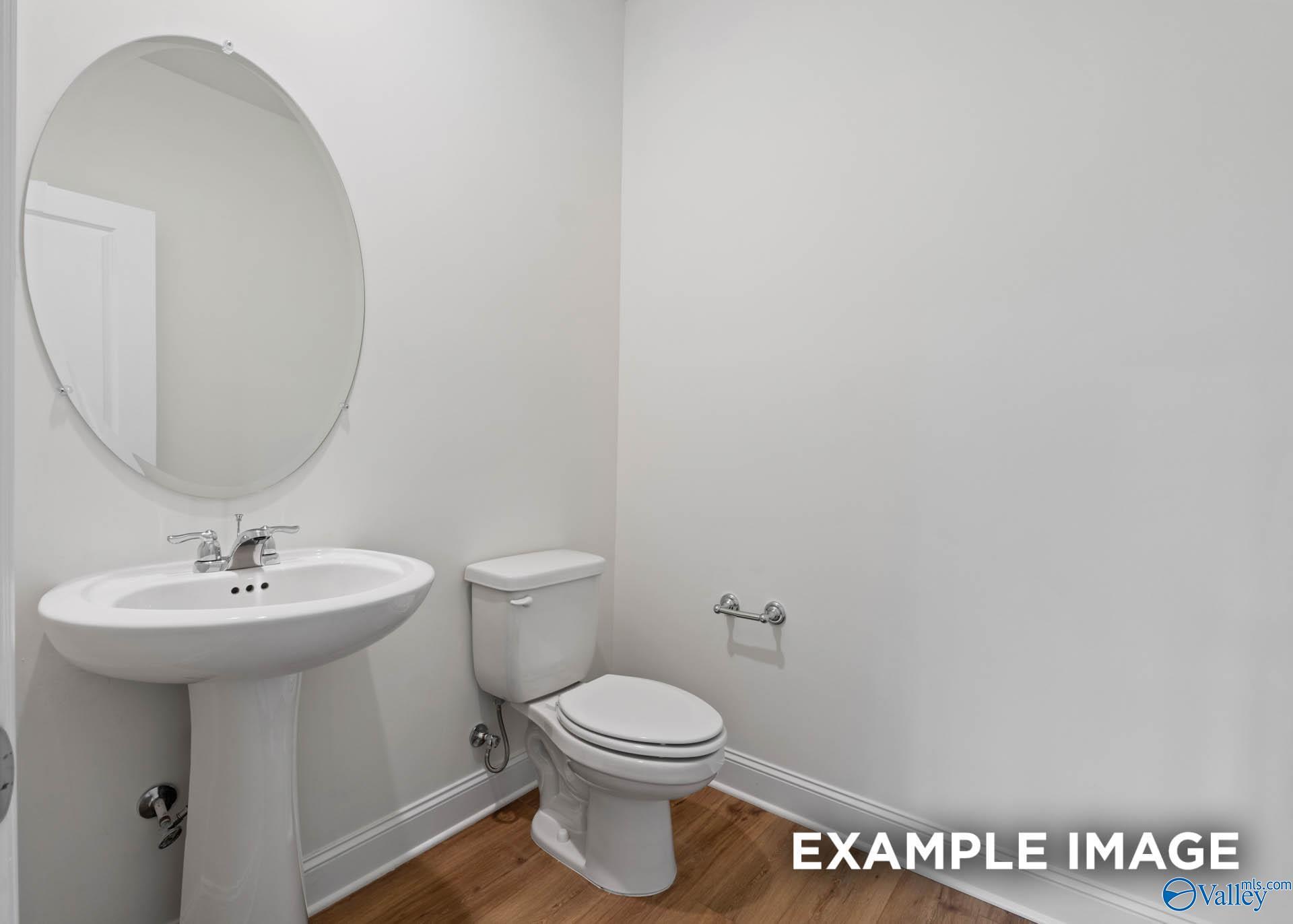 Bright white guest bathroom with pedestal sink, round mirror, and toilet in Evermore Homes The Daphne E, Harvest, Alabama