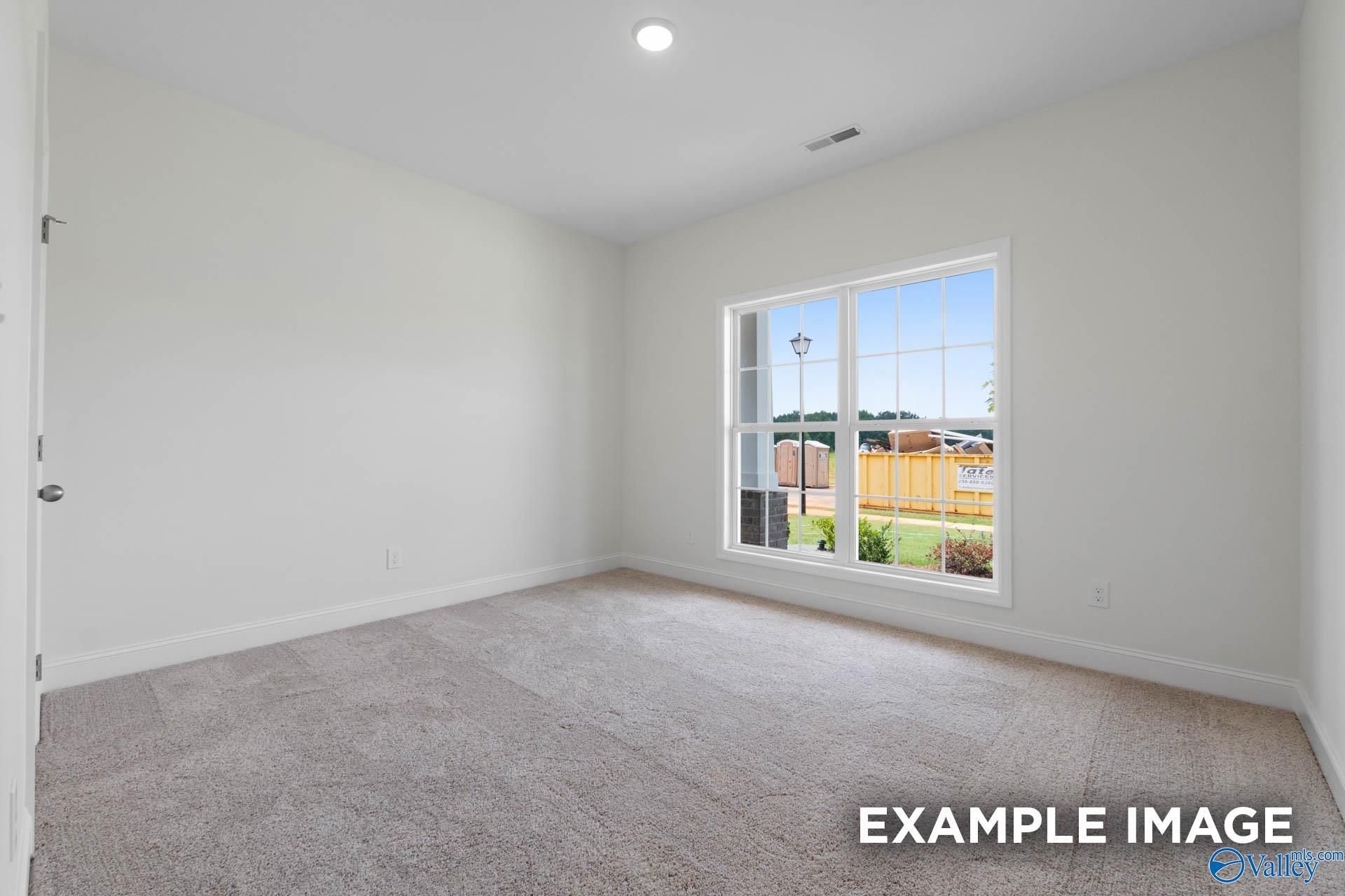 Bright empty bedroom with white walls, beige carpet, and large window overlooking green yard in Evermore Homes Daphne E, Harvest AL