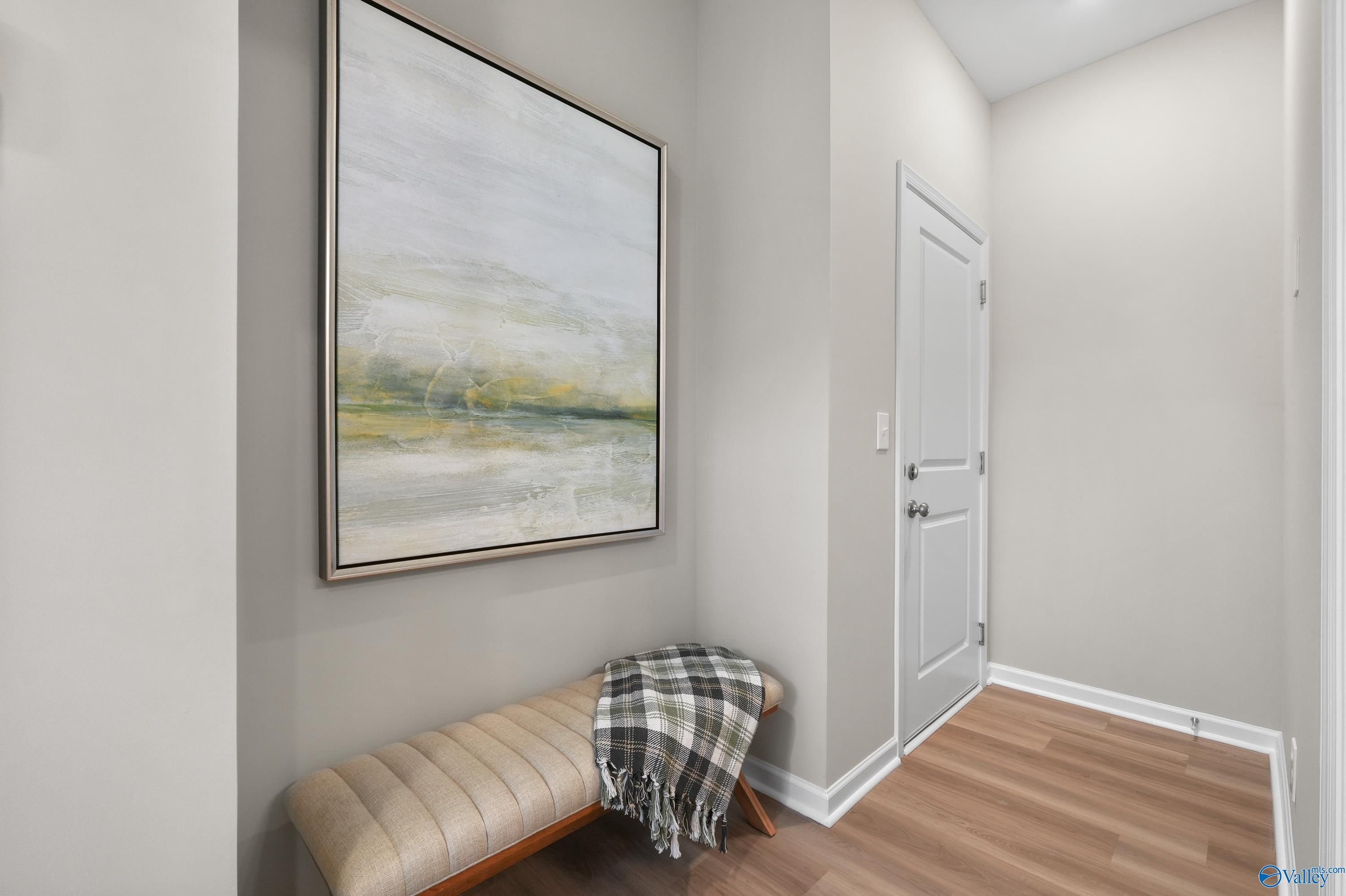 Elegant hallway with abstract landscape painting, plush bench with plaid throw, and light wood floors in Evermore Homes The Malibu, Madison, Alabama