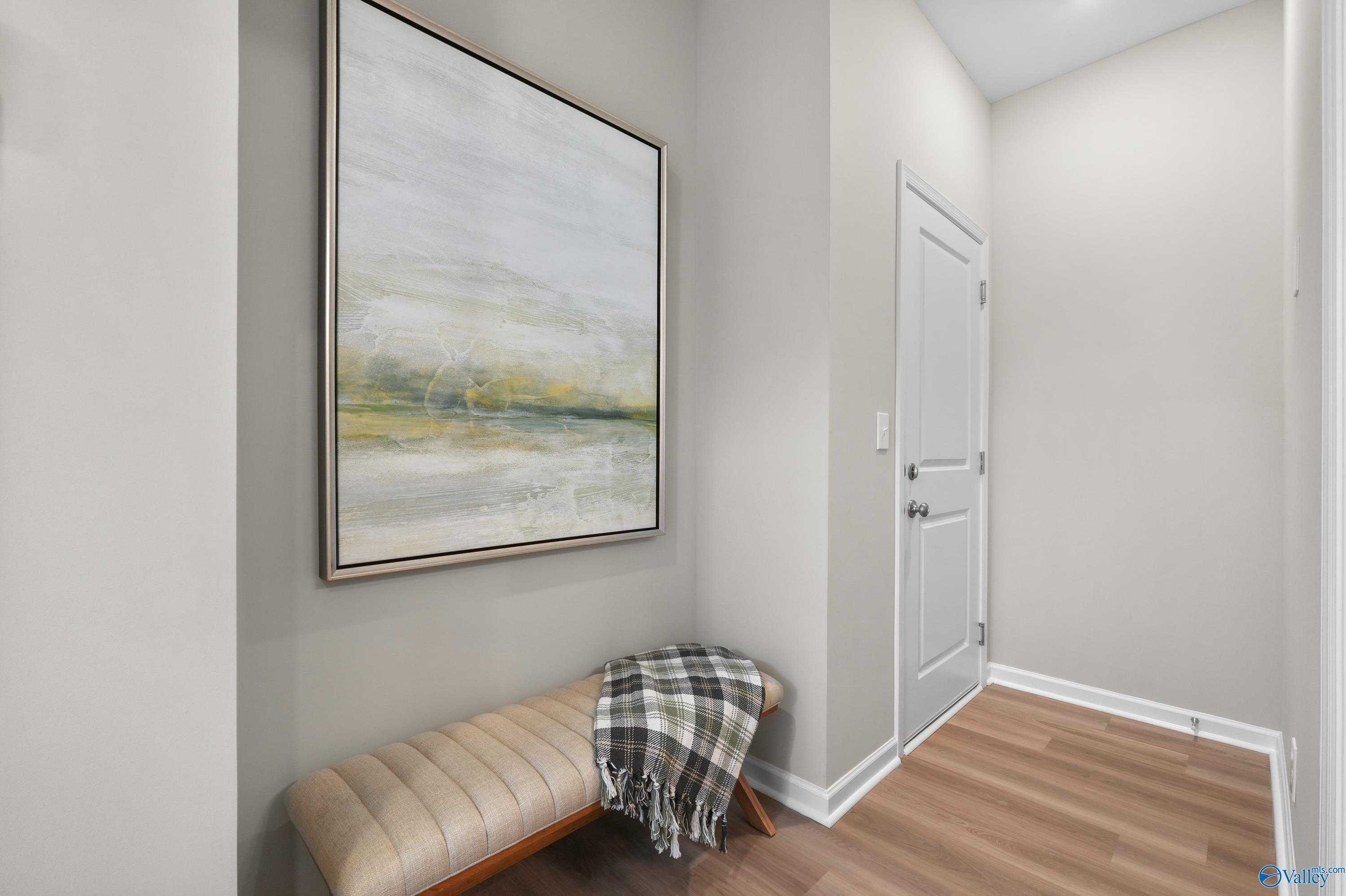 Elegant hallway with abstract landscape painting, plush bench with plaid throw, and light wood floors in Evermore Homes The Malibu, Madison, Alabama