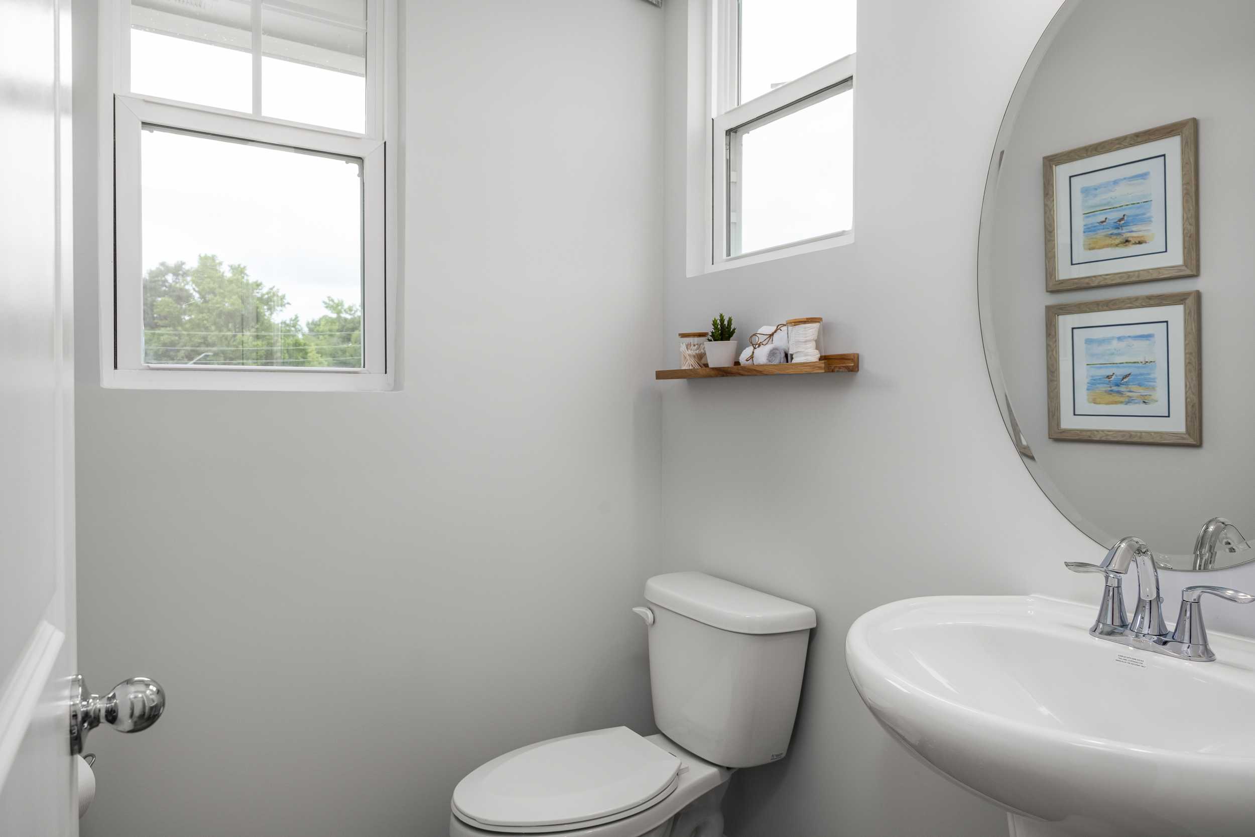 Bright white powder room in Springvale, Fuquay-Varina NC by Davidson Homes with round mirror, wooden shelf plants, and pedestal sink