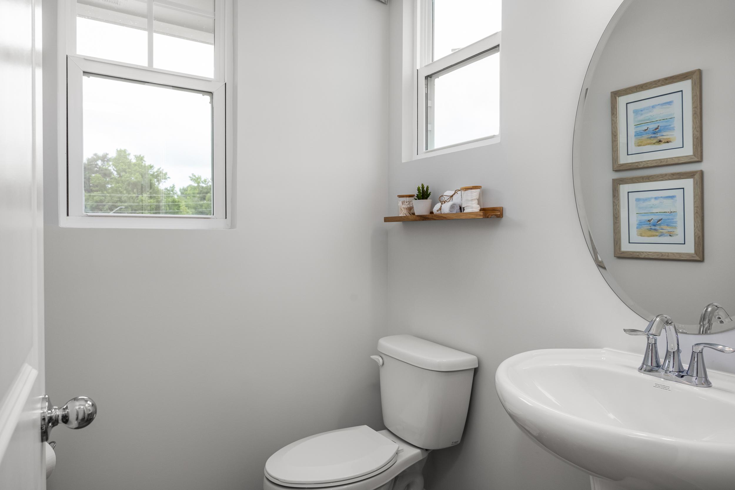 Bright white powder room in Springvale, Fuquay-Varina NC by Davidson Homes with round mirror, wooden shelf plants, and pedestal sink