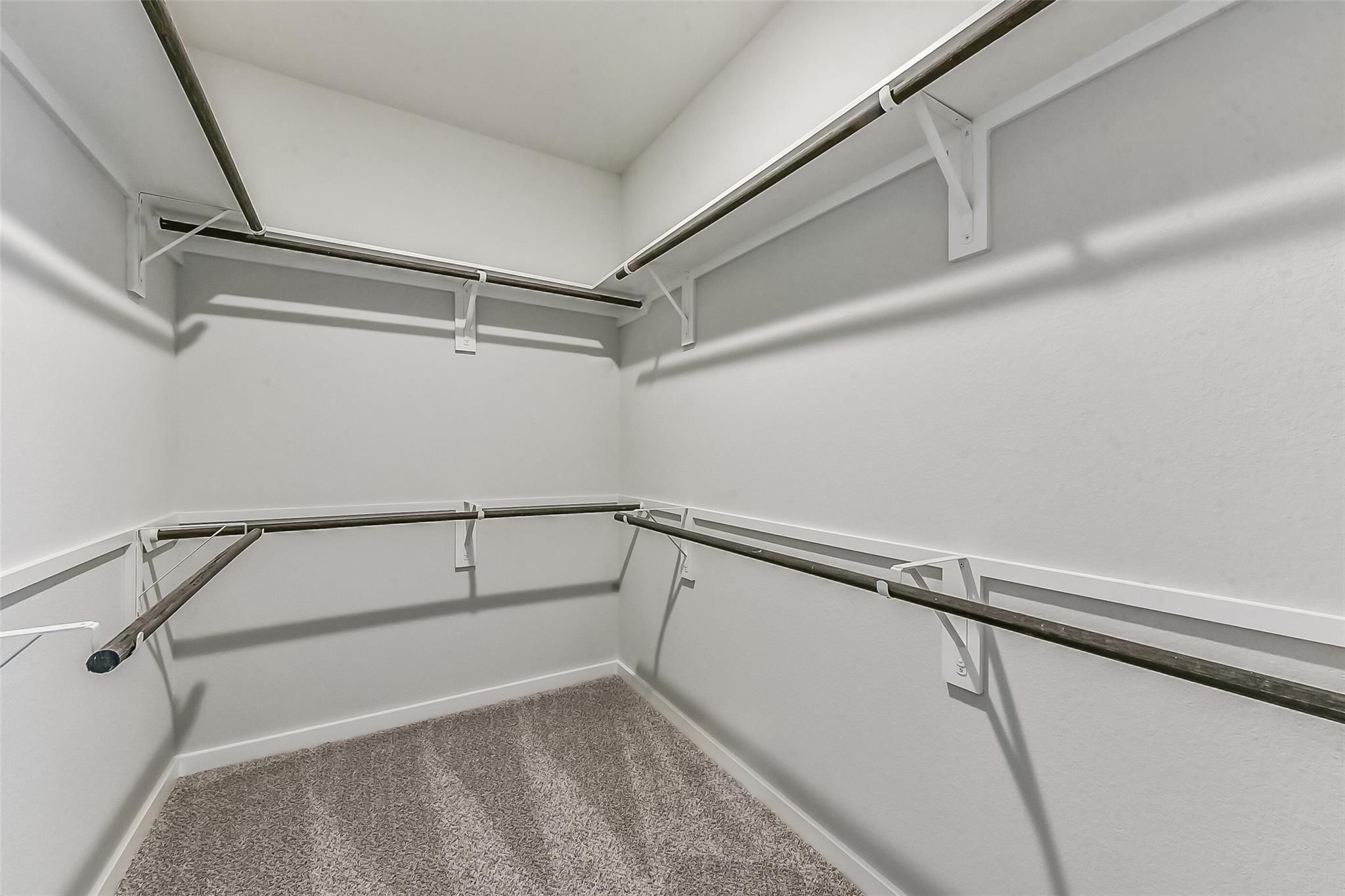 Spacious L-shaped walk-in closet with metal hanging rods, white walls, and beige carpet in Davidson Homes The Costa B, Dayton, Texas