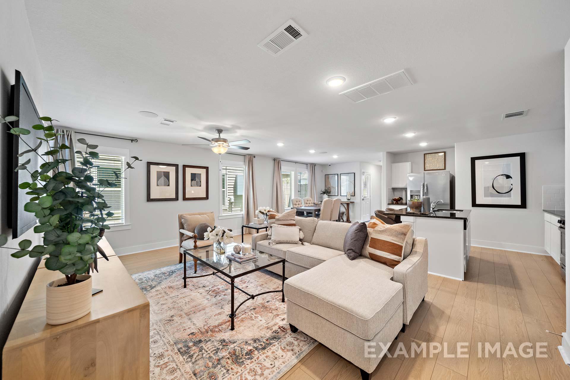 Spacious open-concept living room in The Rose B with beige L-shaped sofa, glass coffee table, potted plant, and adjacent white kitchen island