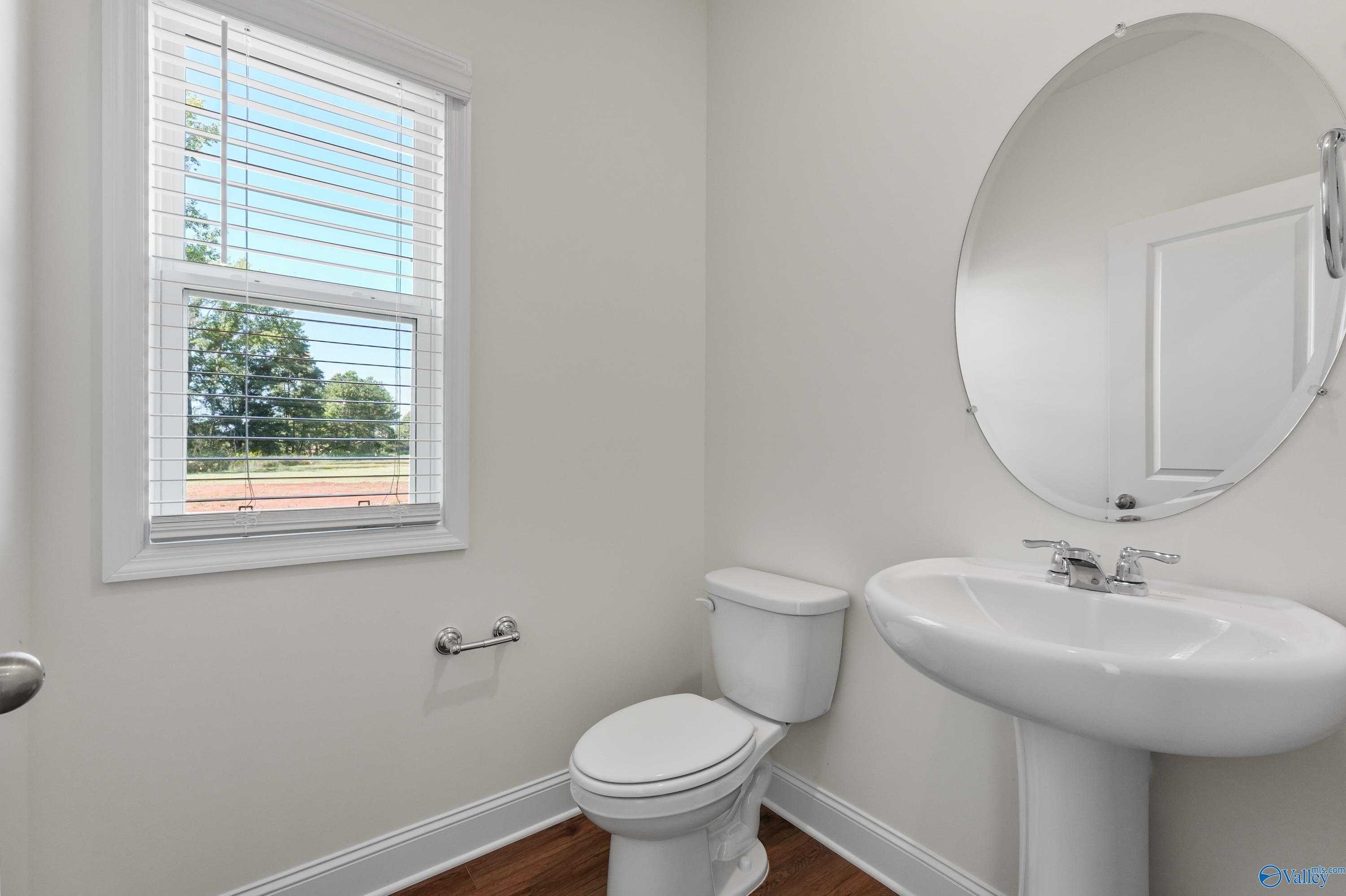 Elegant powder room featuring white pedestal sink, round mirror, toilet, and window view in Davidson Homes The Shelby A, Athens, Alabama
