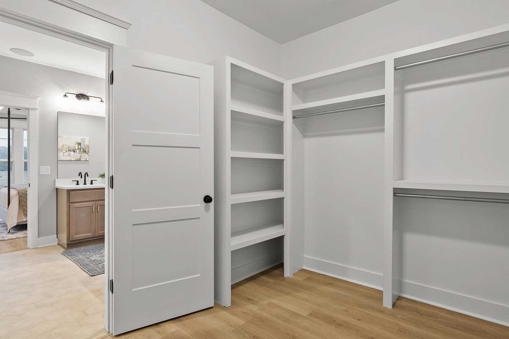 Spacious The Oxford master walk-in closet with built-in white shelves, rods, open doors to adjacent bathroom vanity